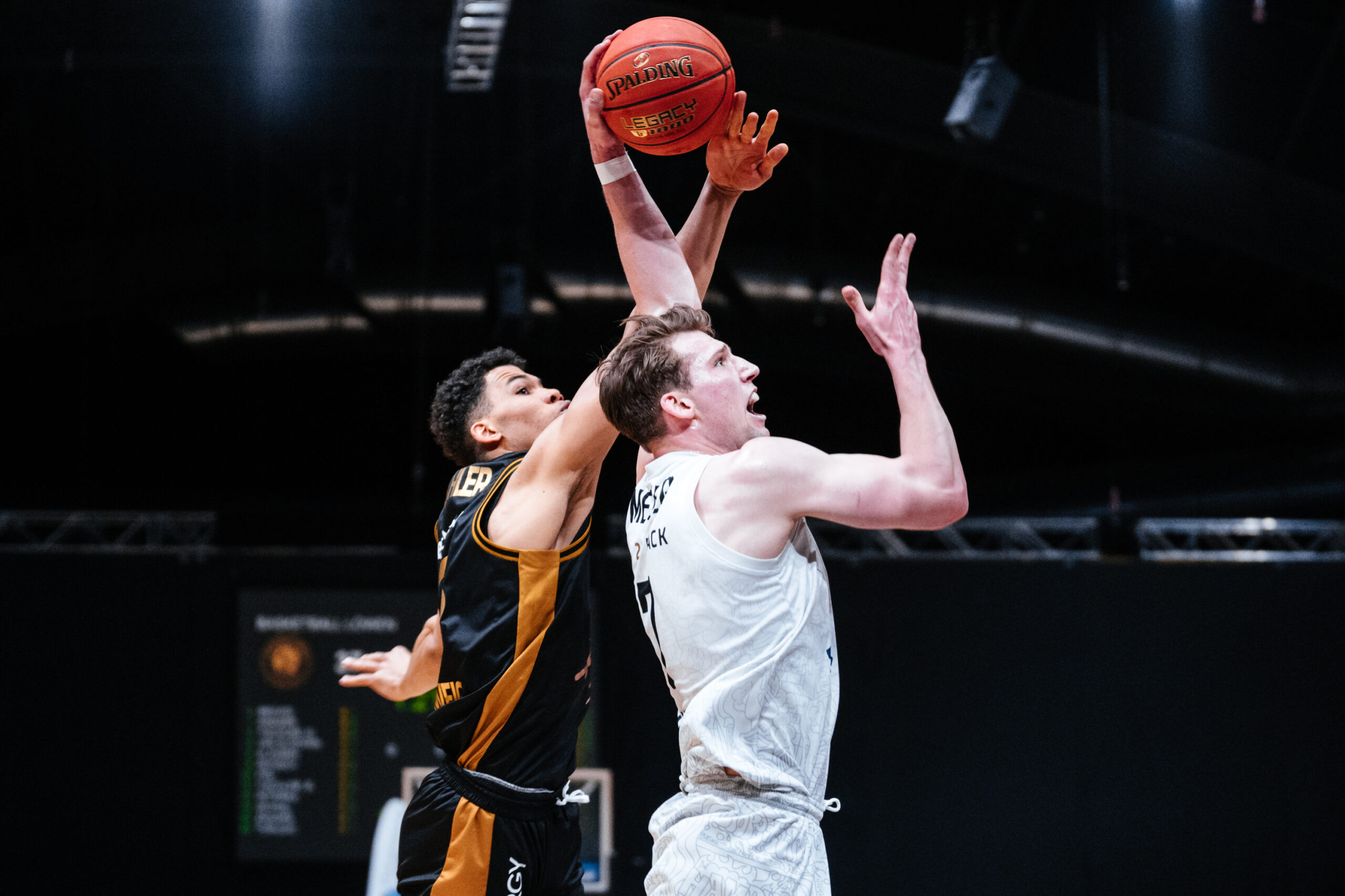 Zwei Basketballspieler im Sprung, einer in weiß versucht zu werfen, der andere in schwarz-gelb blockt den Ball