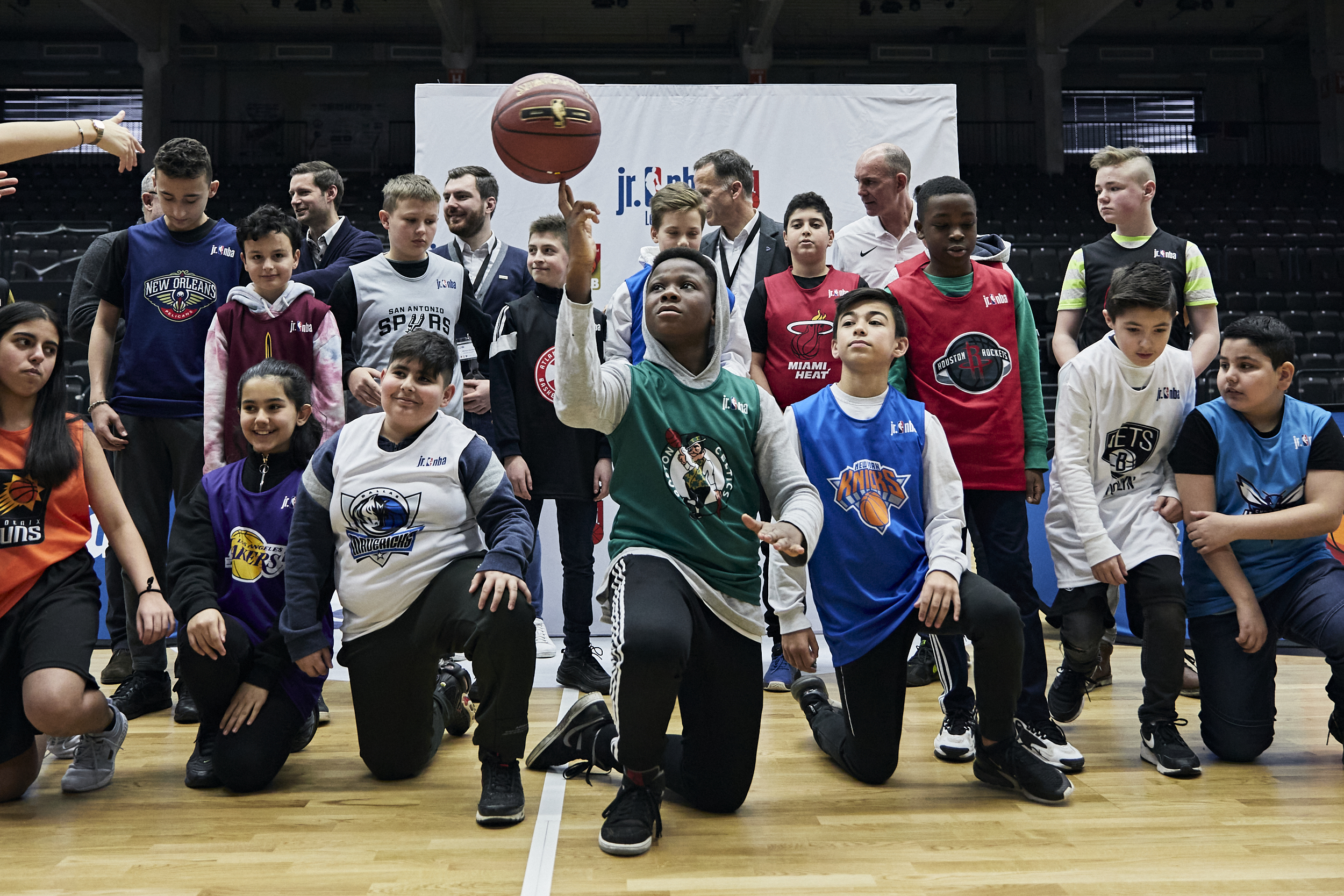 Gruppe von Kindern in Basketballtrikots verschiedener Teams, einige knien auf dem Hallenboden, ein Junge wirft einen Basketball in die Luft