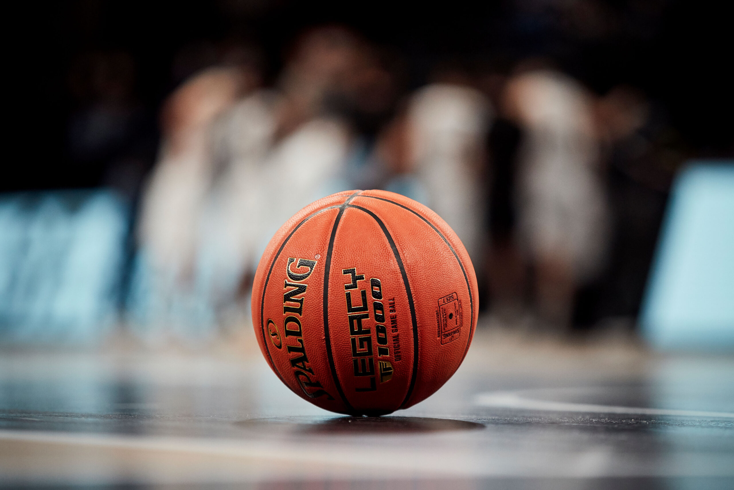 Oranger Basketball mit der Aufschrift 'Spalding Legacy' auf dem Spielfeld, unscharfe Spieler im Hintergrund