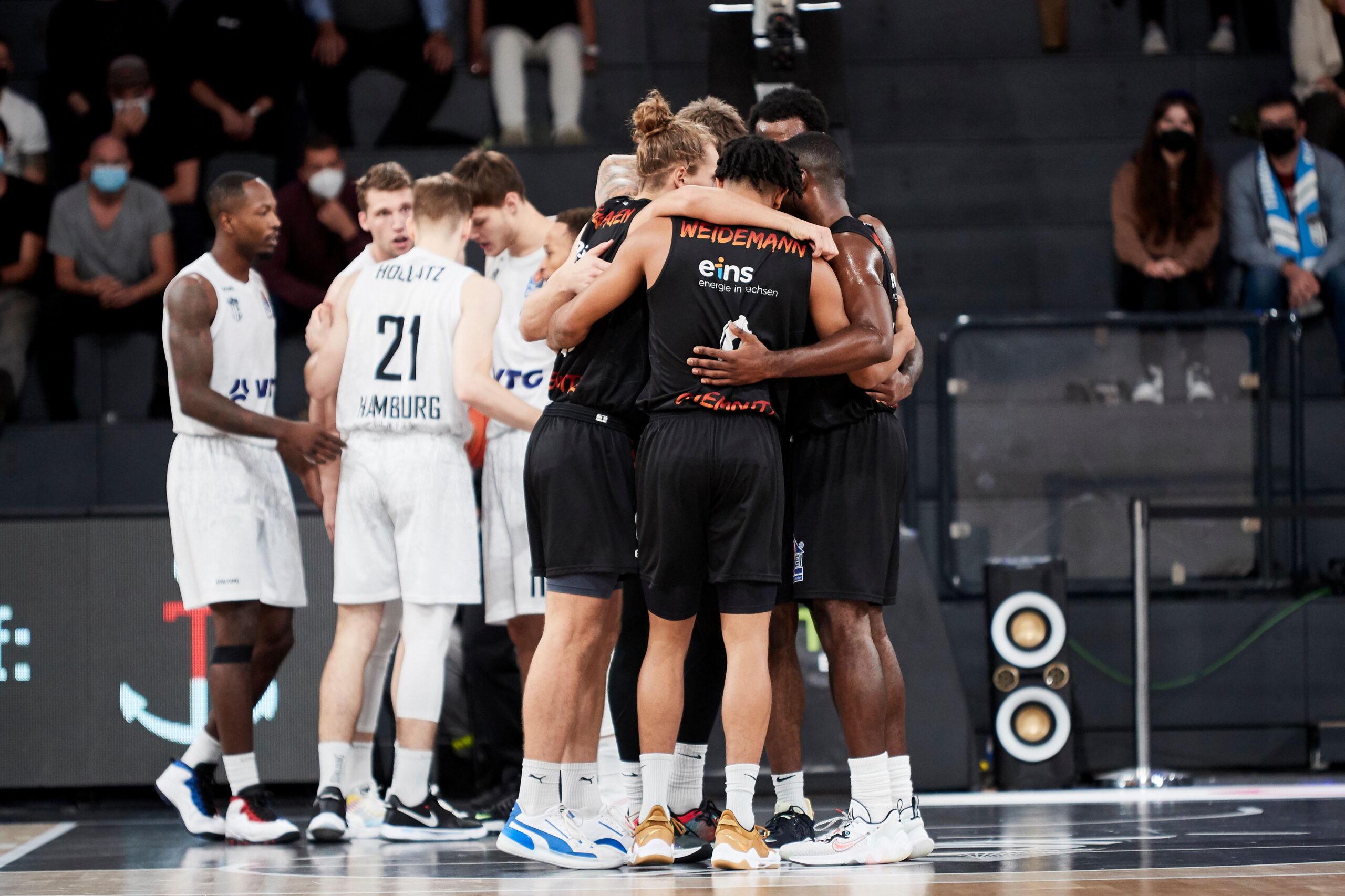 Basketballspieler in schwarzen Trikots umarmen sich im Kreis, während Spieler in weißen Trikots im Hintergrund stehen