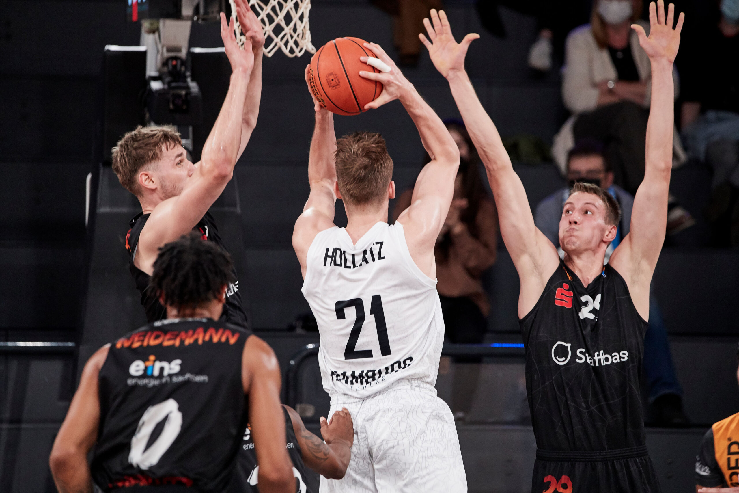 Basketballspieler mit Trikotnummer 21 wirft Korb, zwei Verteidiger mit erhobenen Armen versuchen zu blocken