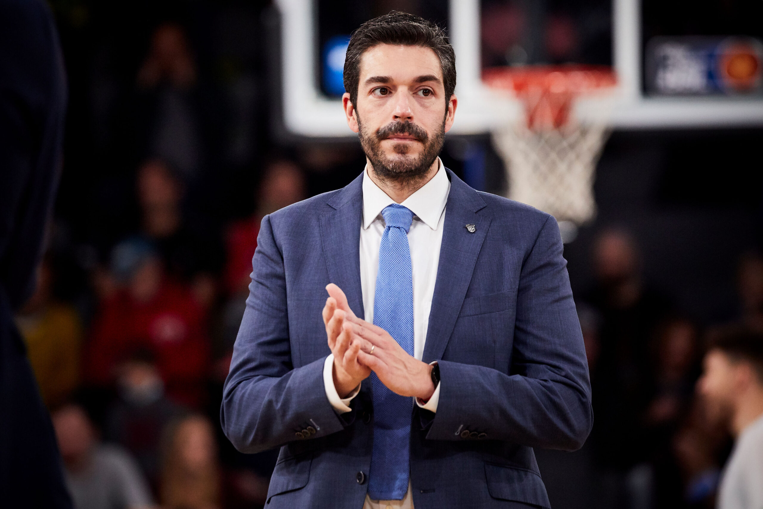 Mann in blauem Anzug und hellblauer Krawatte klatscht vor einem Basketballkorb im Hintergrund