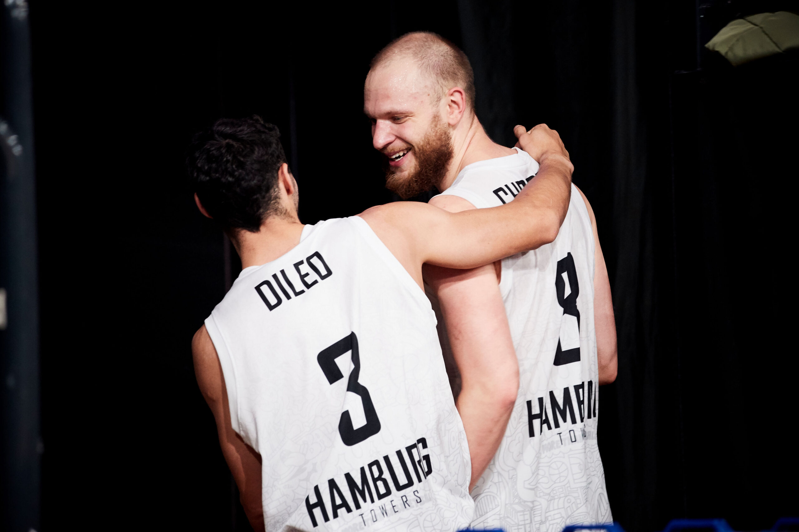 Zwei Basketballspieler der Hamburg Towers umarmen sich, tragen weiße Trikots mit den Nummern 3 und 9