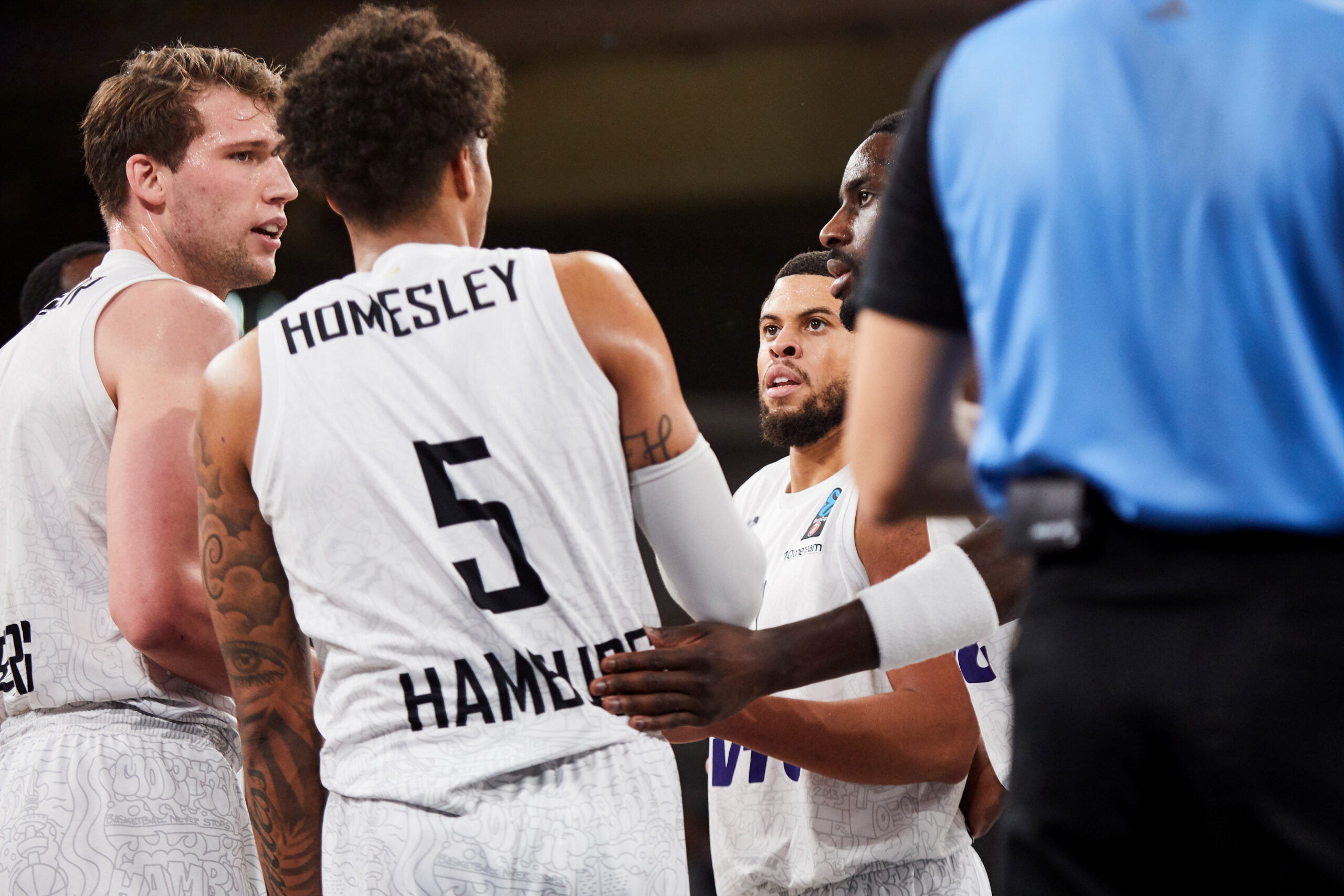 Basketballspieler in weißen Trikots mit der Aufschrift 'Homesley Hamburg' während einer Spielpause, Schiedsrichter im Vordergrund