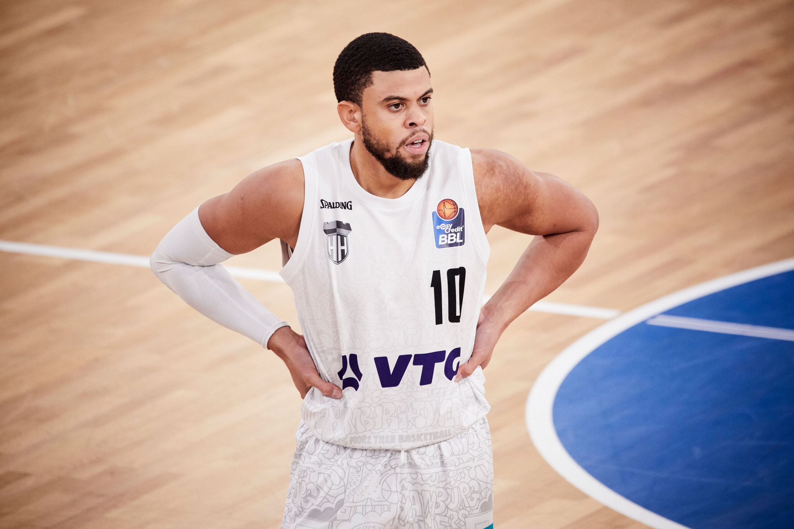 Basketballspieler in weißem Trikot mit der Nummer 10 steht mit Händen in der Hüfte auf dem Spielfeld