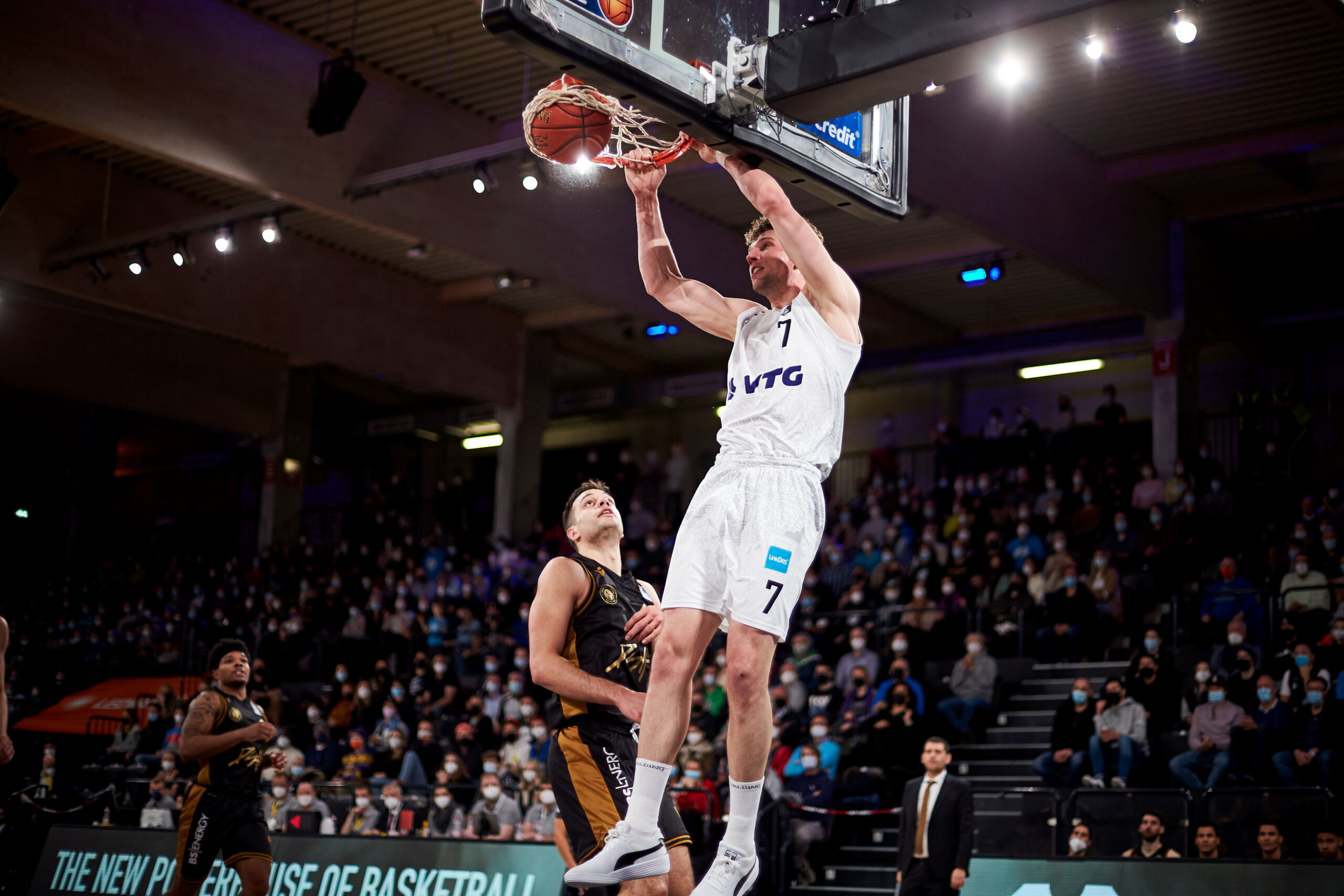 Basketballspieler in weißem Trikot mit der Nummer 7 macht einen Dunk vor einem verteidigenden Spieler in schwarzem Trikot, Zuschauer im Hintergrund