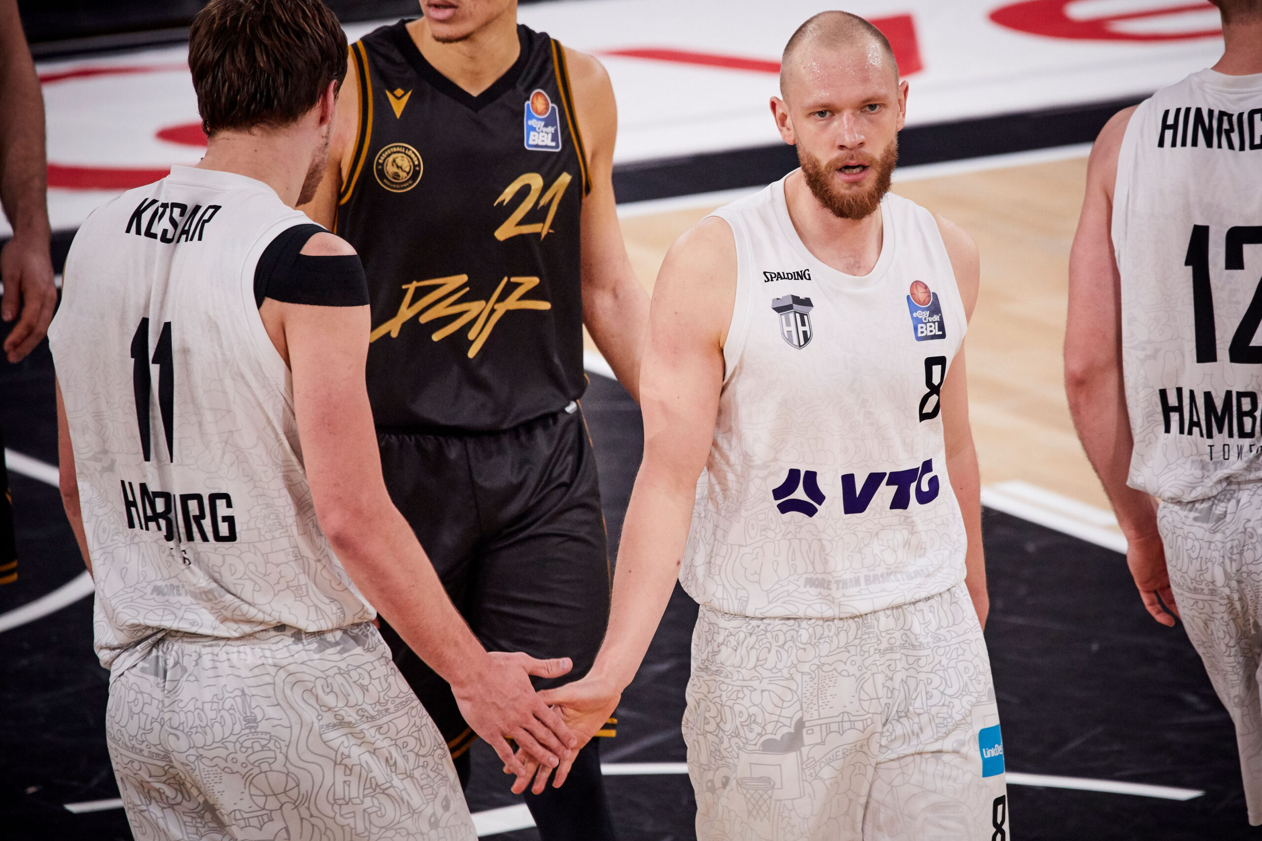Zwei Basketballspieler in weißen Trikots mit den Nummern 11 und 8 geben sich die Hand auf dem Spielfeld während eines Spiels.