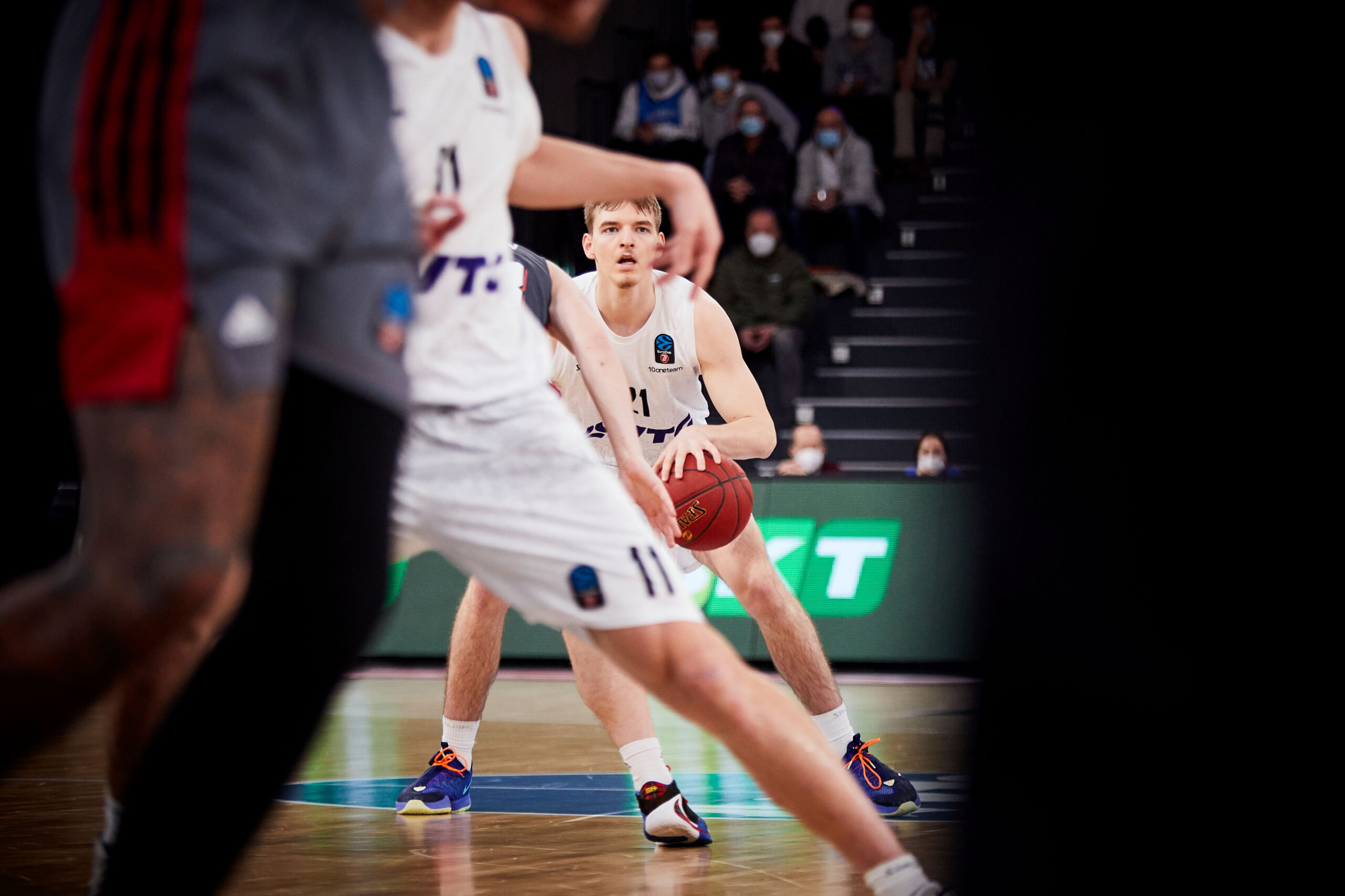 Basketballspieler in weißem Trikot mit Ball in der Hand, fokussiert auf das Spiel, umgeben von Gegnern auf dem Spielfeld