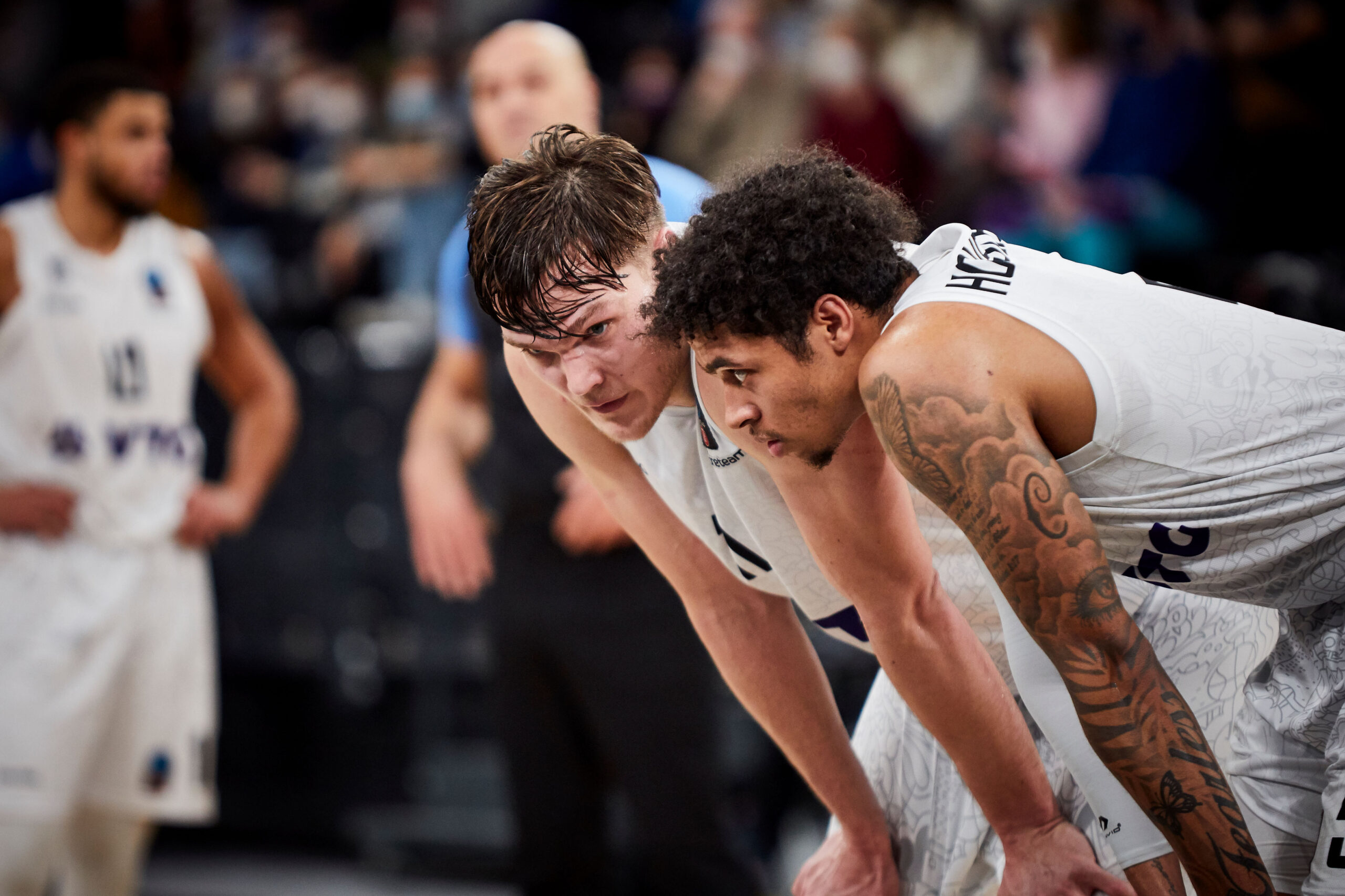 Zwei Basketballspieler in weißen Trikots beugen sich erschöpft nach vorne, im Hintergrund weitere Spieler und Zuschauer unscharf