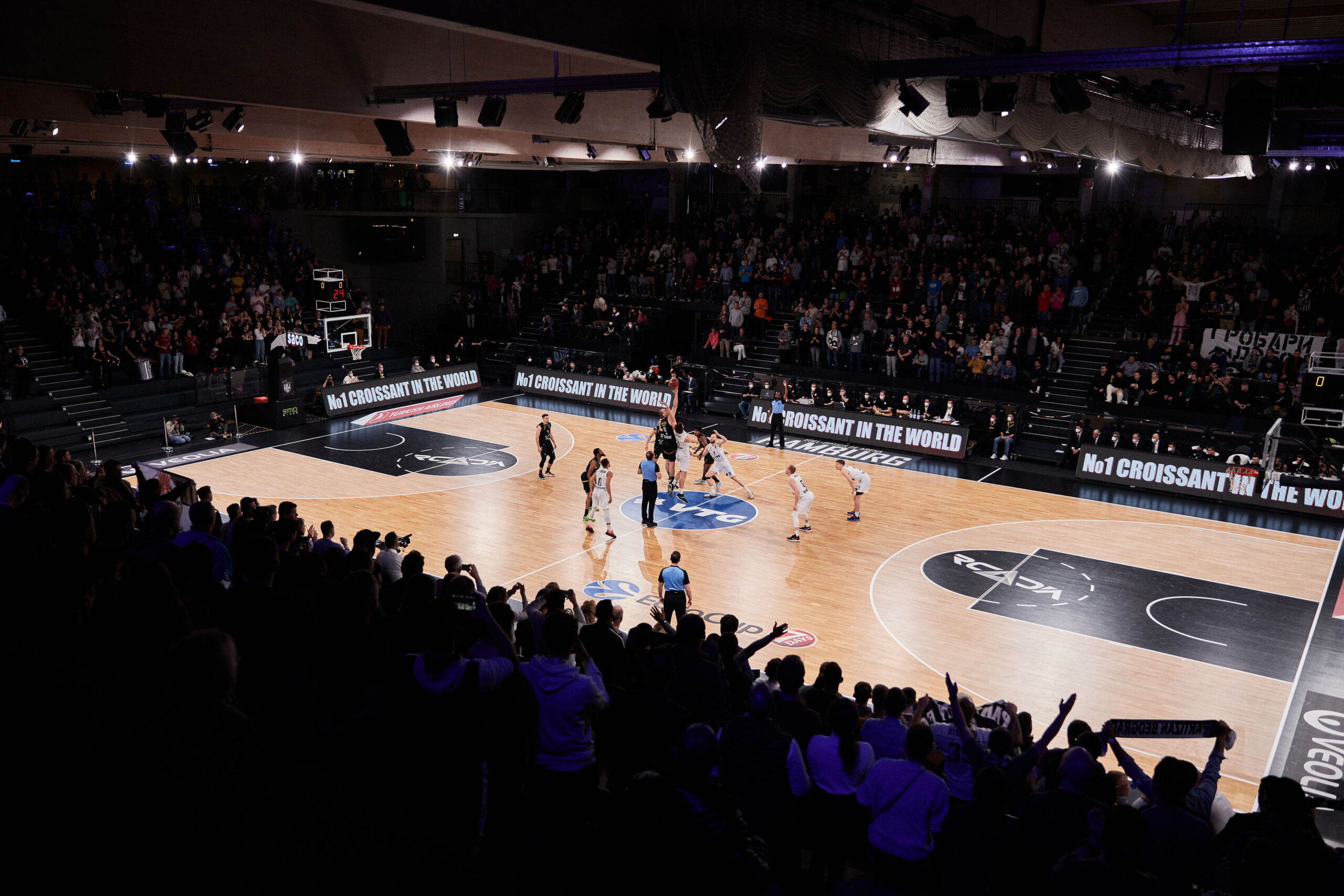 Basketballspiel in einer Halle mit Zuschauern auf den Rängen und Spielern auf dem Spielfeld