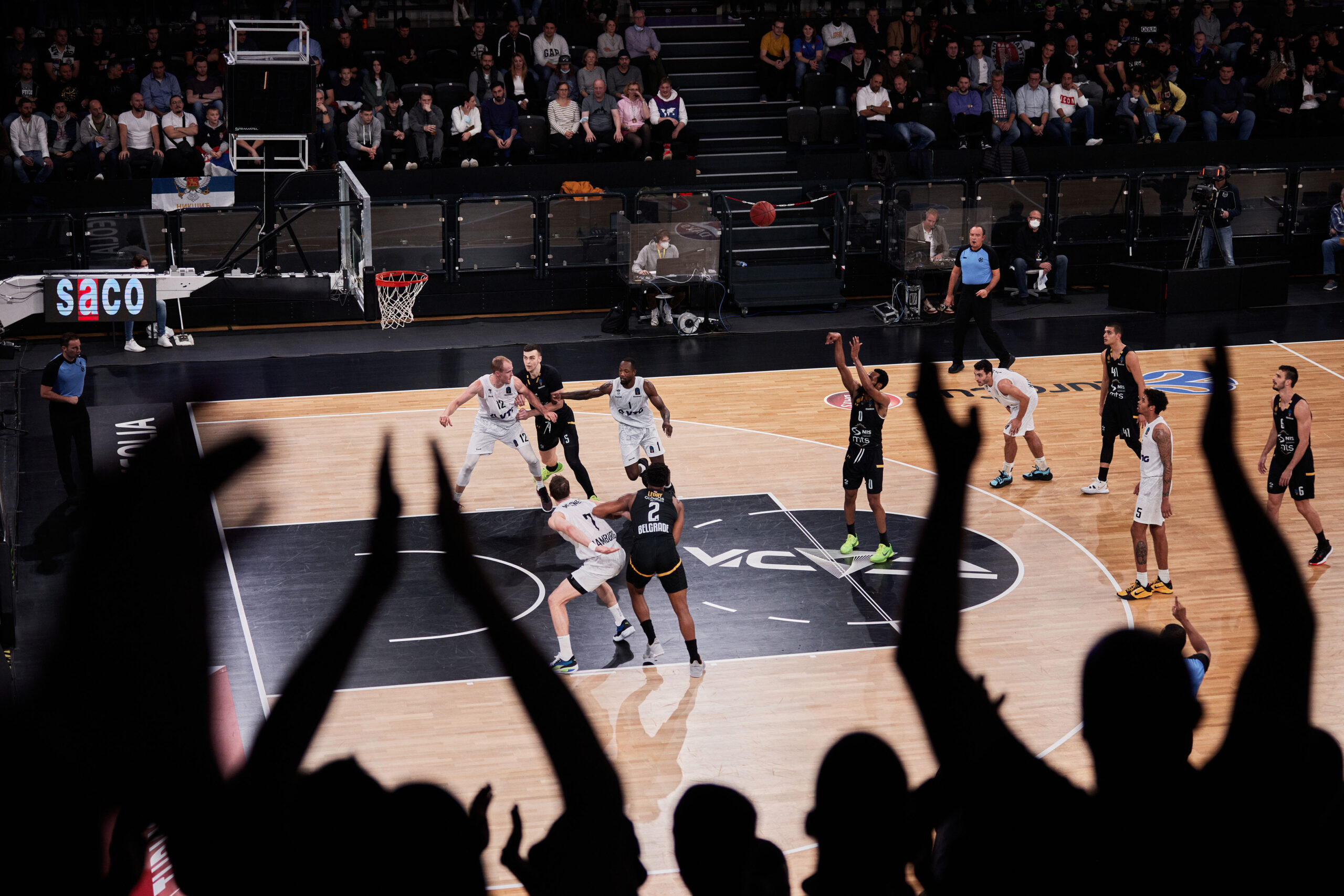 Basketballspiel in einer Halle mit jubelnden Zuschauern im Vordergrund, Spieler in weißen und schwarzen Trikots auf dem Spielfeld