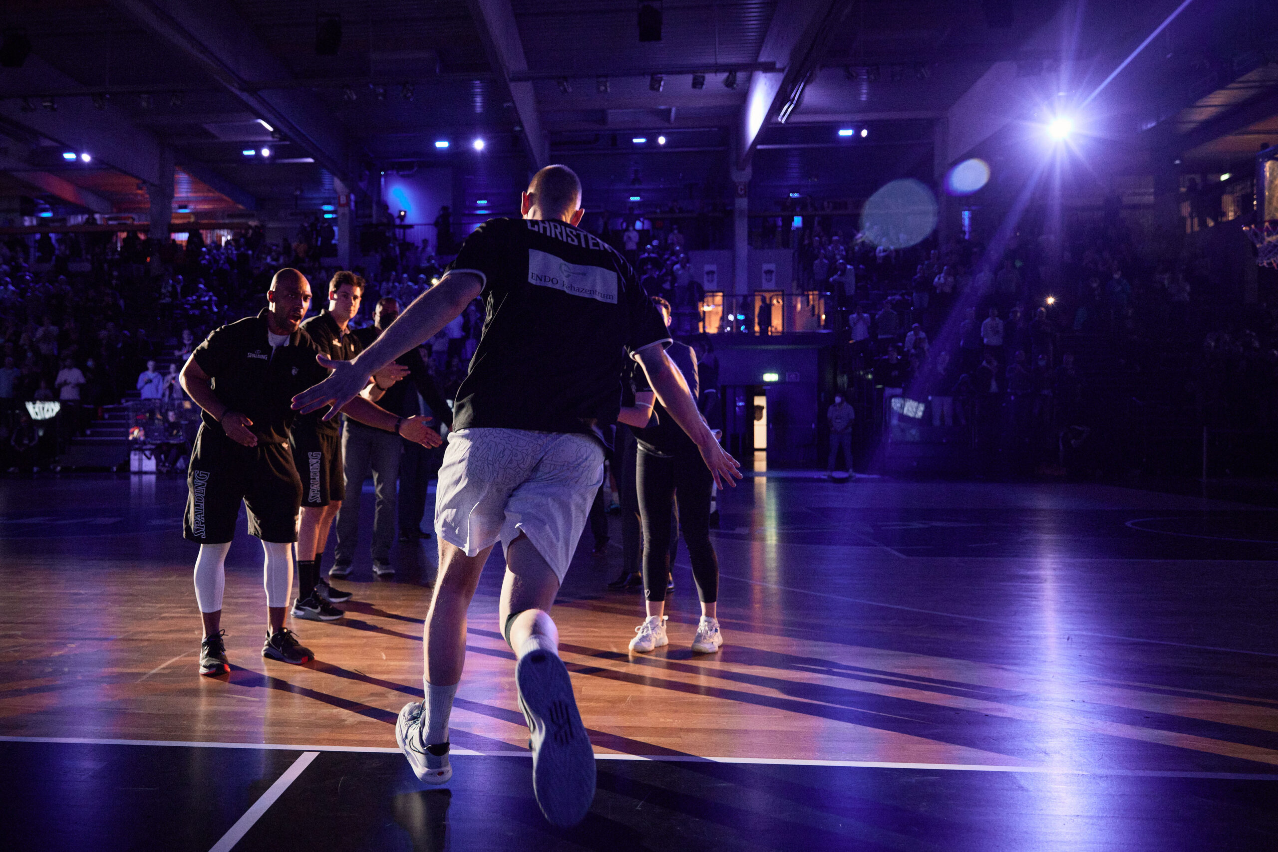 Basketballspieler in Sportkleidung betritt Spielfeld, umgeben von Teamkollegen und Publikum in einer Halle mit Bühnenbeleuchtung