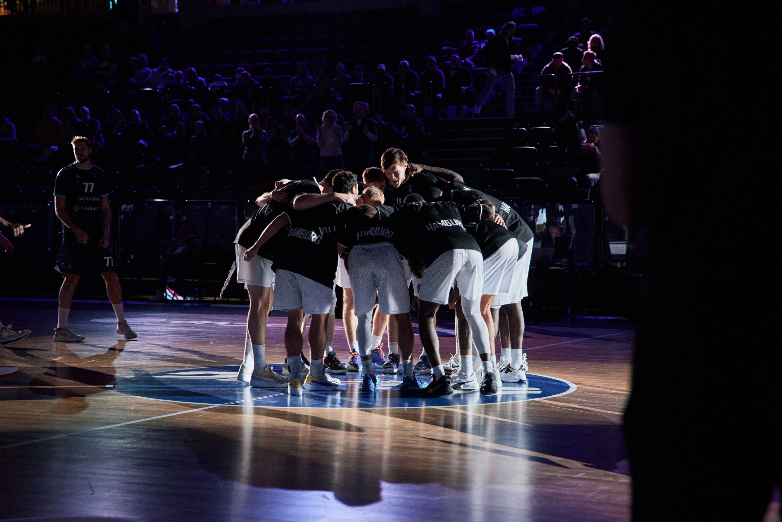 Basketballteam in schwarzer Kleidung und weißen Shorts steht im Kreis auf dem Spielfeld, um sich vor dem Spiel zu motivieren