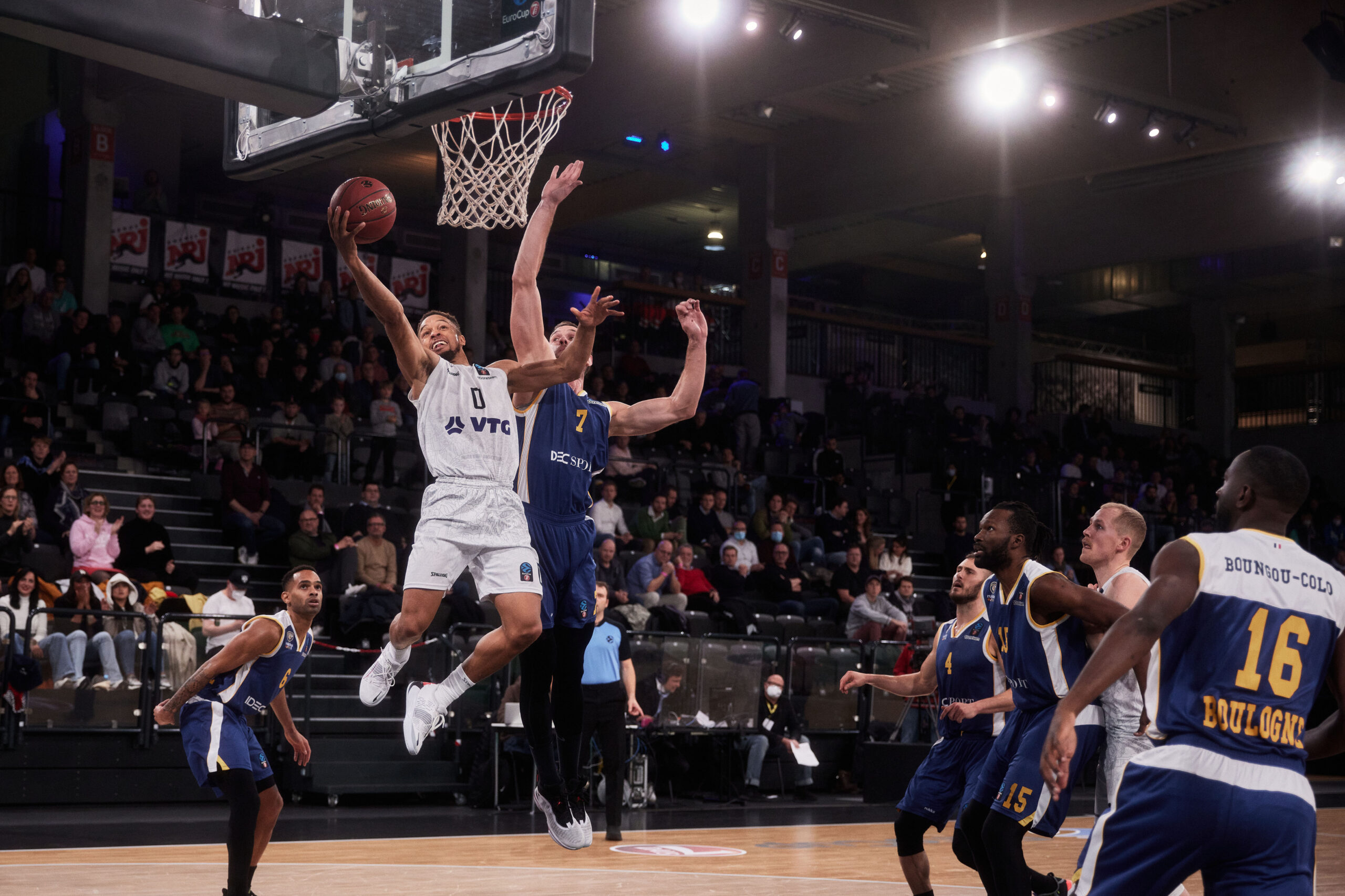 Basketballspieler in weißer Uniform springt zum Korb, um einen Wurf zu machen, während Spieler in blauen Trikots verteidigen, Zuschauer im Hintergrund