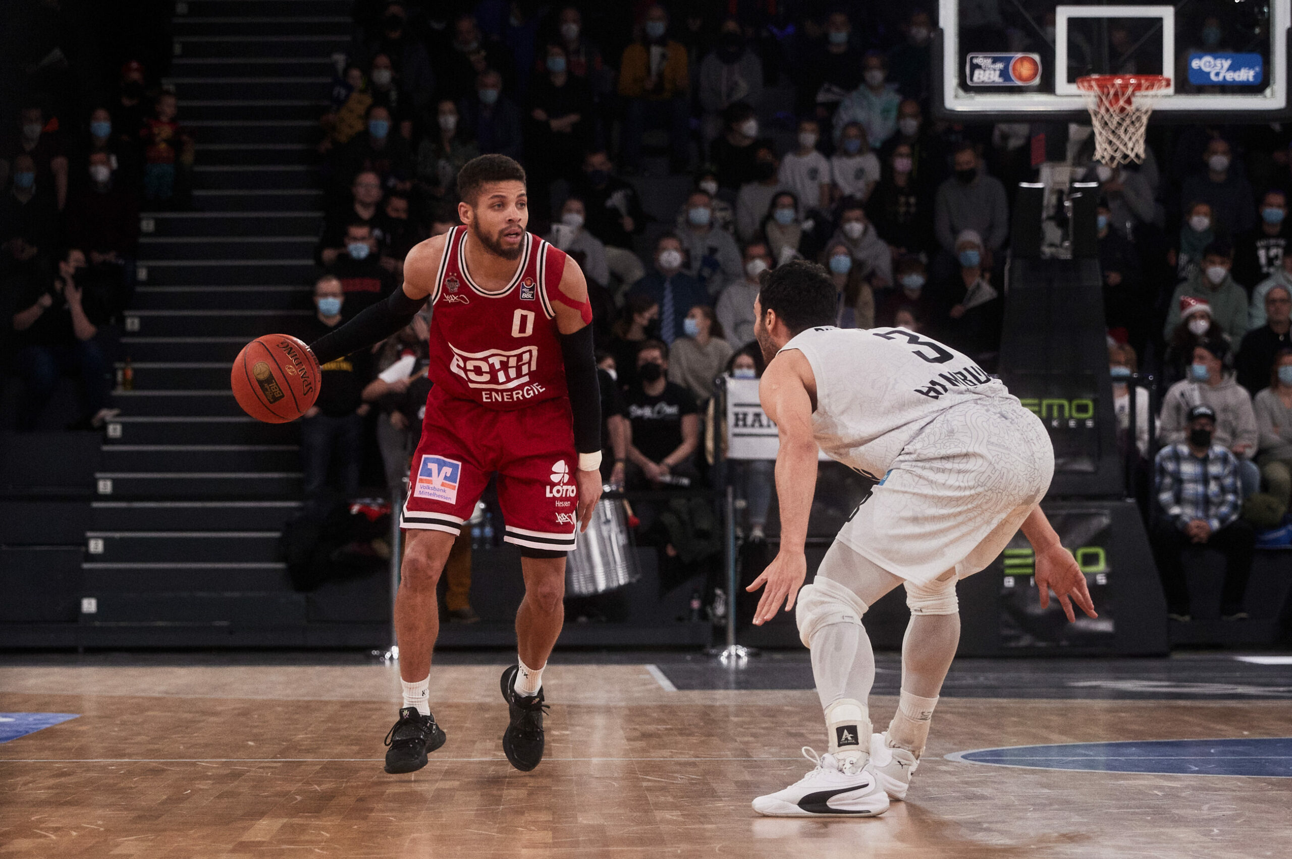 Basketballspieler in rotem Trikot mit Ball wird von einem Spieler in weißem Trikot verteidigt, Zuschauer im Hintergrund