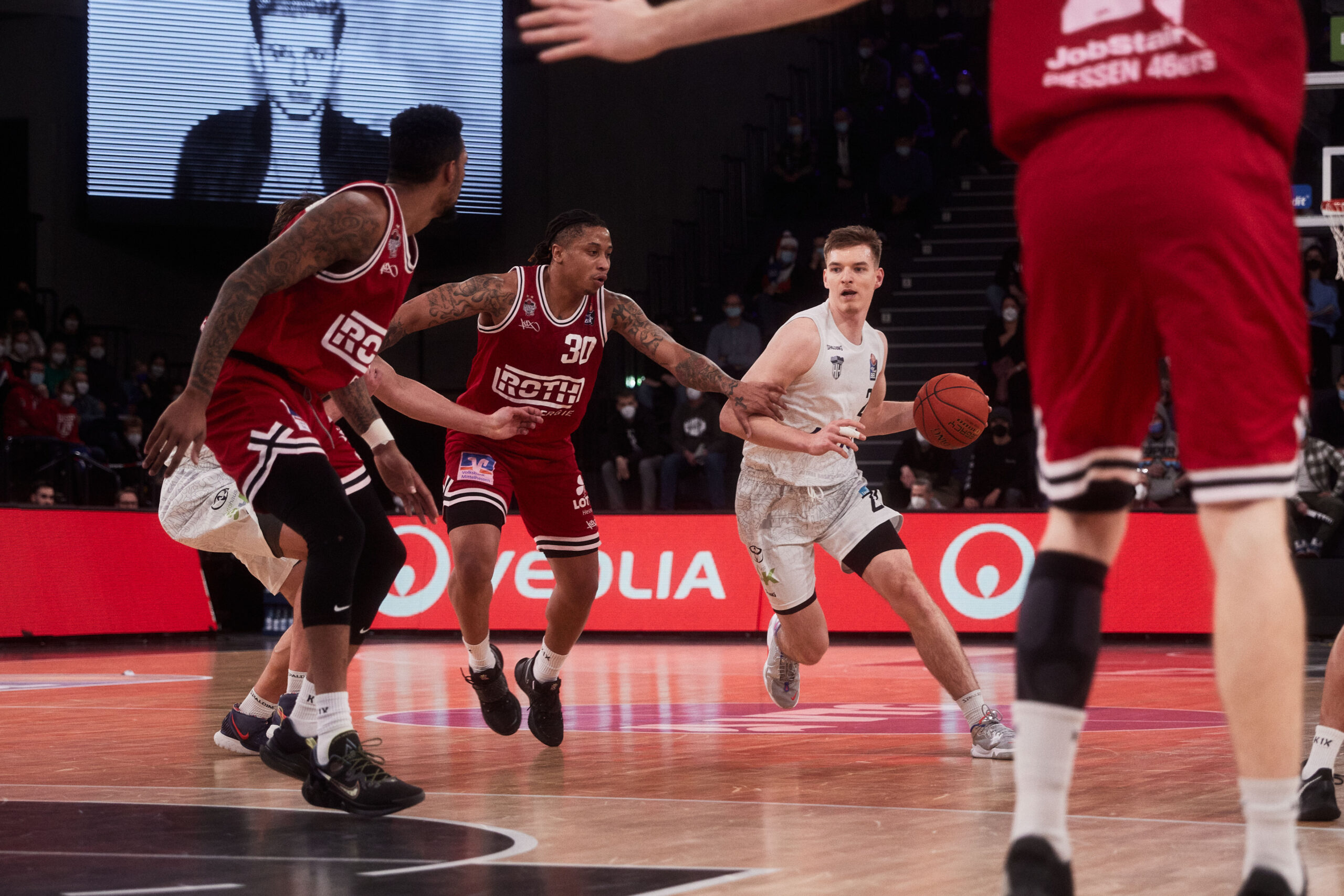 Basketballspieler in weißer Trikot dribbelt den Ball, während drei Spieler in roten Trikots ihn verteidigen auf dem Spielfeld