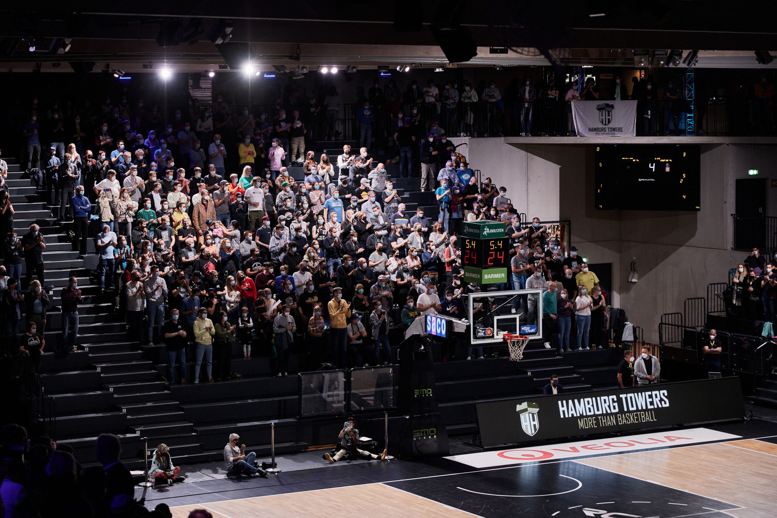 Zuschauer in einer Sporthalle verfolgen ein Basketballspiel, sichtbar ist der Korb und die Anzeigetafel, Banner mit Hamburg Towers Logo