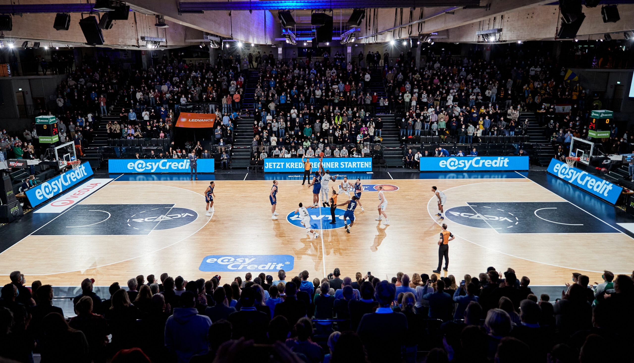 Basketballspiel in einer Halle mit vielen Zuschauern, Spieler beim Sprungball in der Spielfeldmitte