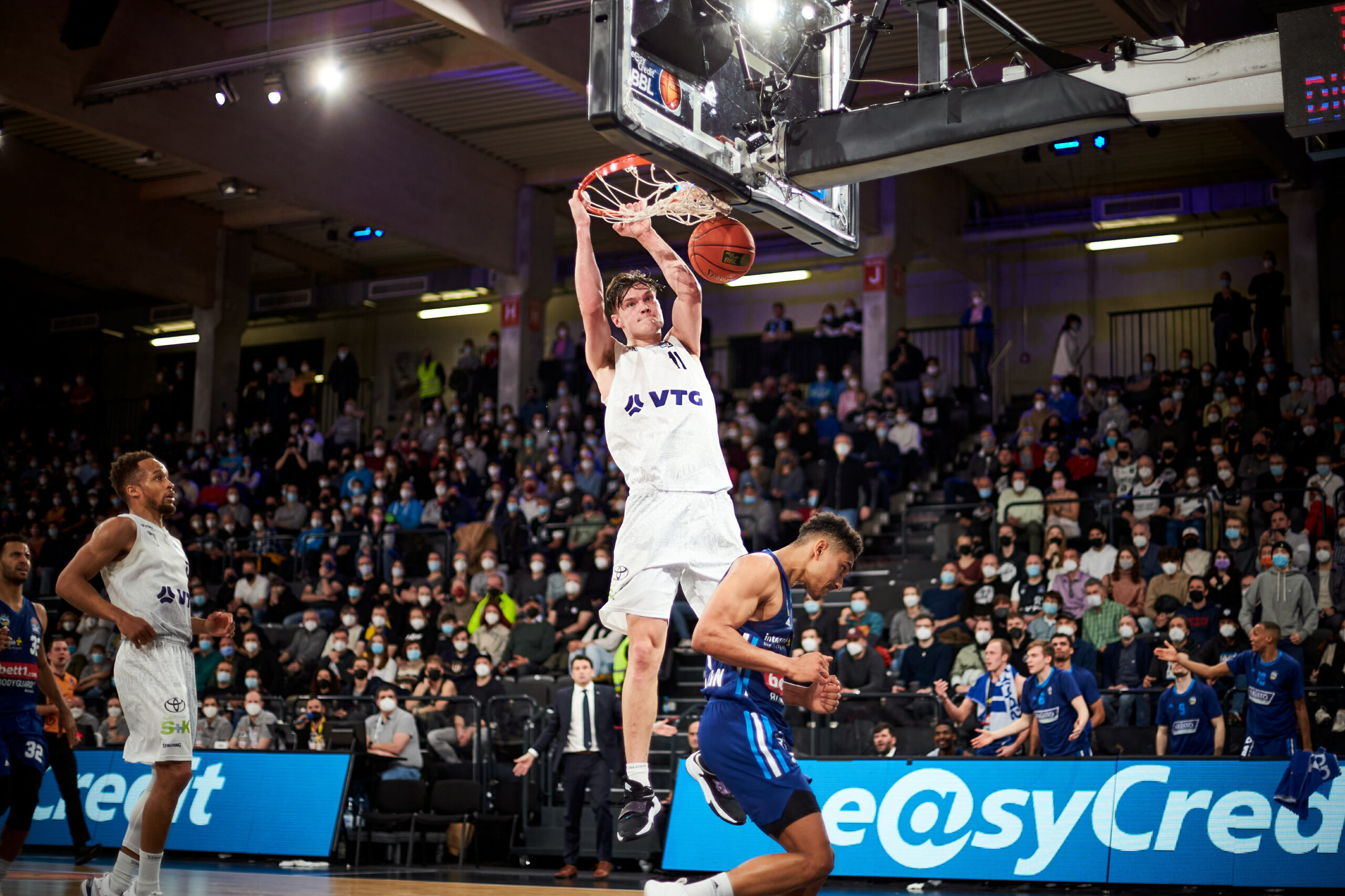 Basketballspieler in weißem Trikot macht Dunking vor Publikum in Halle