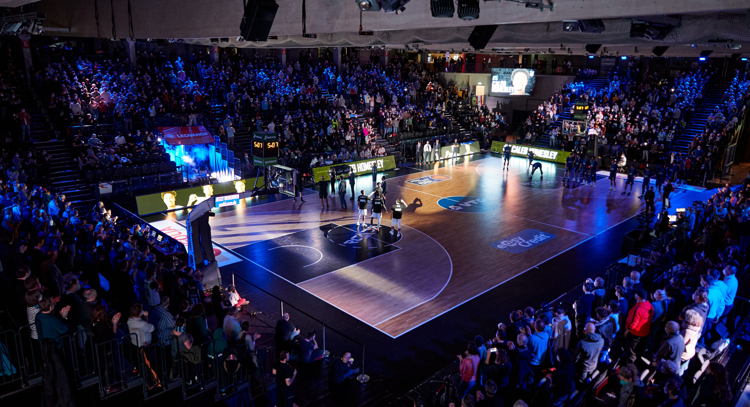 Basketballspiel in einer Halle mit vielen Zuschauern, Spielfeld beleuchtet, Spieler und Schiedsrichter auf dem Feld