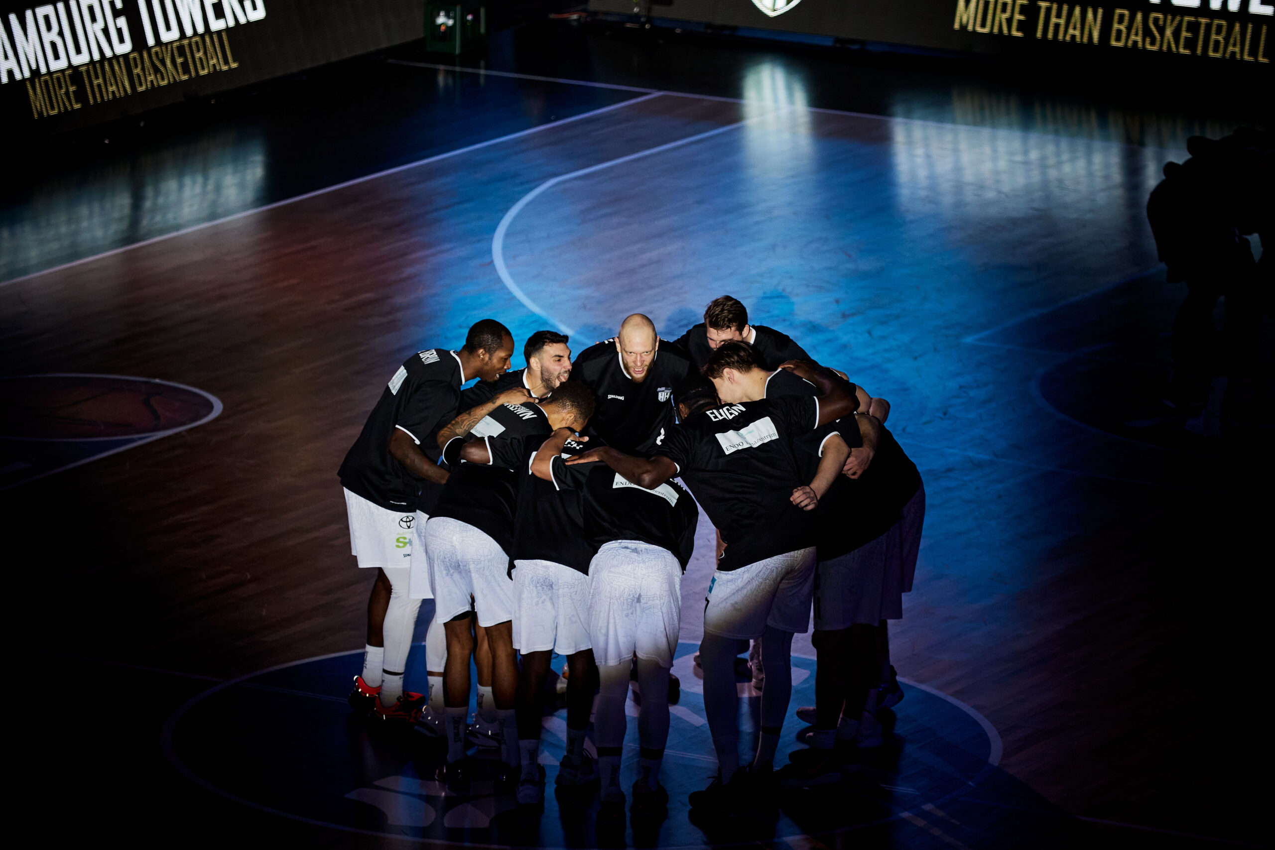 Basketballteam in schwarzer Trainingskleidung steht im Kreis auf dem Spielfeld und bespricht sich