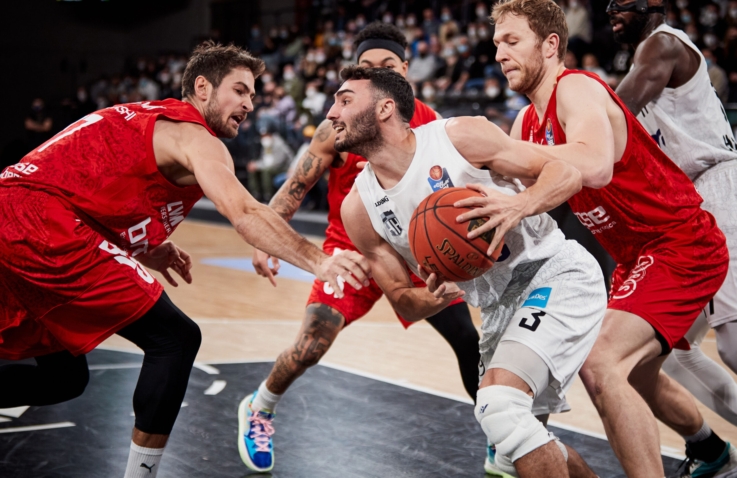 Basketballspieler in weißem Trikot dribbelt den Ball, umgeben von drei Gegnern in roten Trikots, die versuchen, ihn zu blockieren.