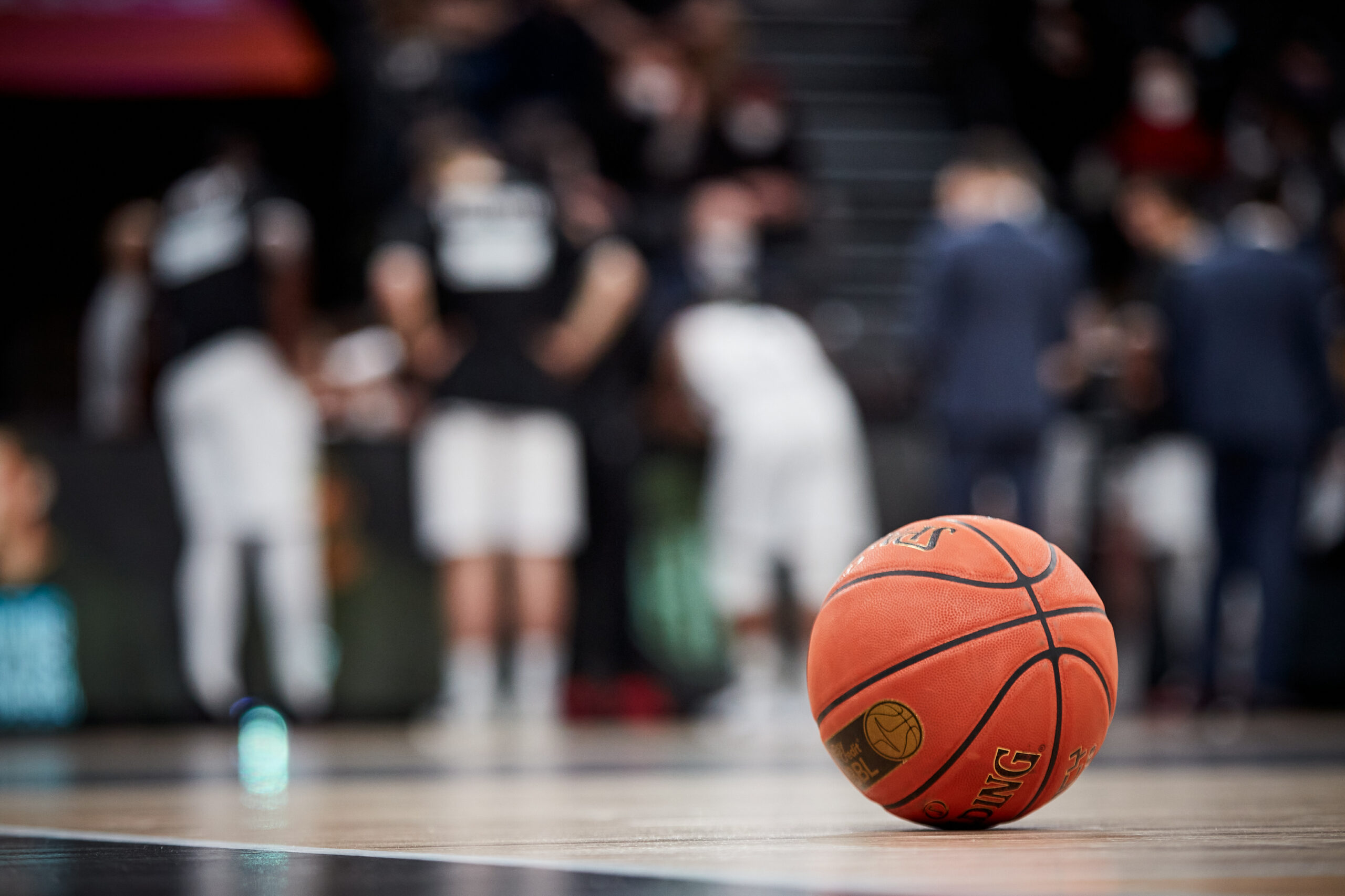 Basketball liegt auf dem Spielfeld, im Hintergrund unscharfe Spieler und Zuschauer