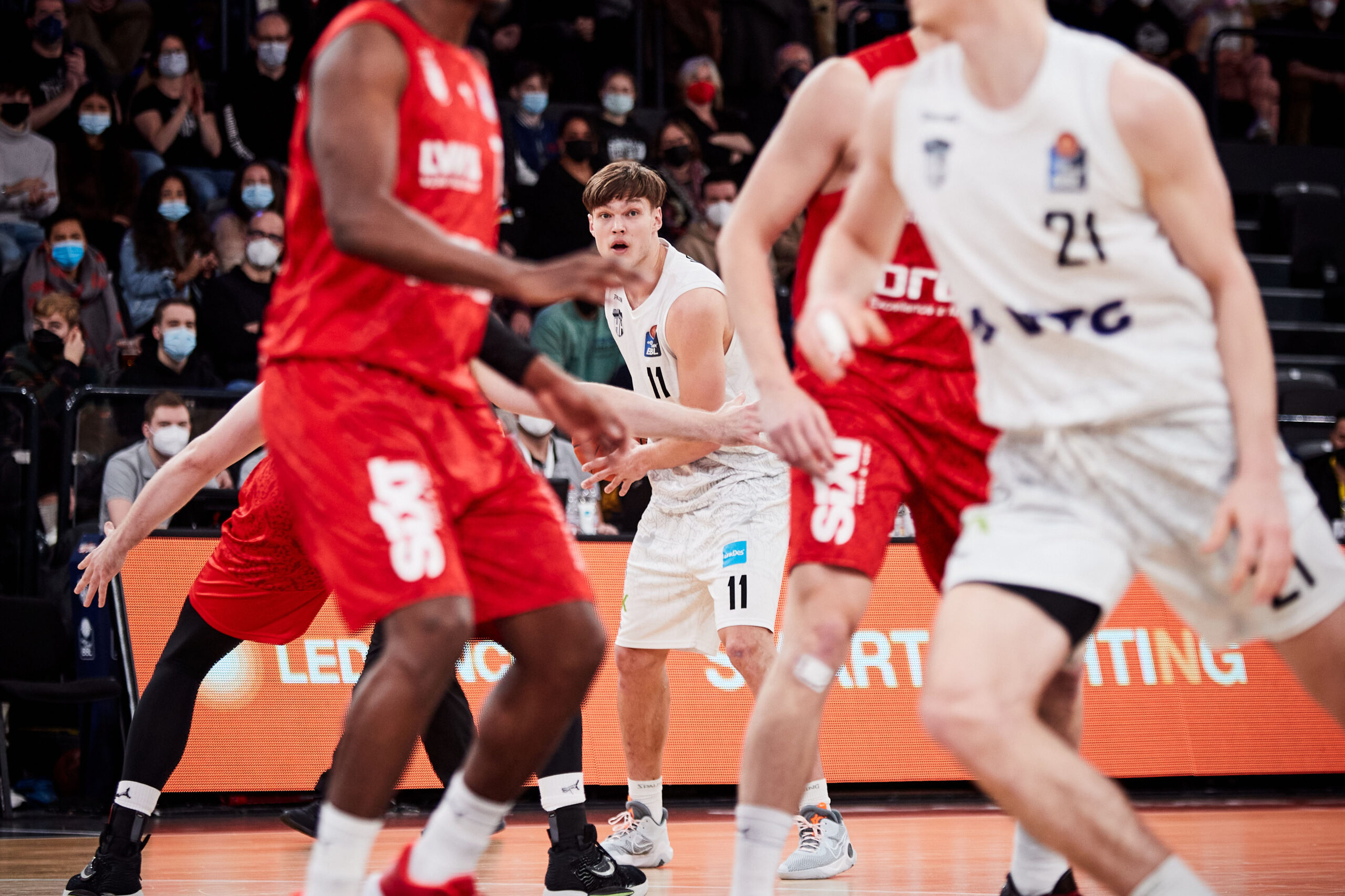 Basketballspieler in weißen und roten Trikots während eines Spiels, Zuschauer mit Masken im Hintergrund