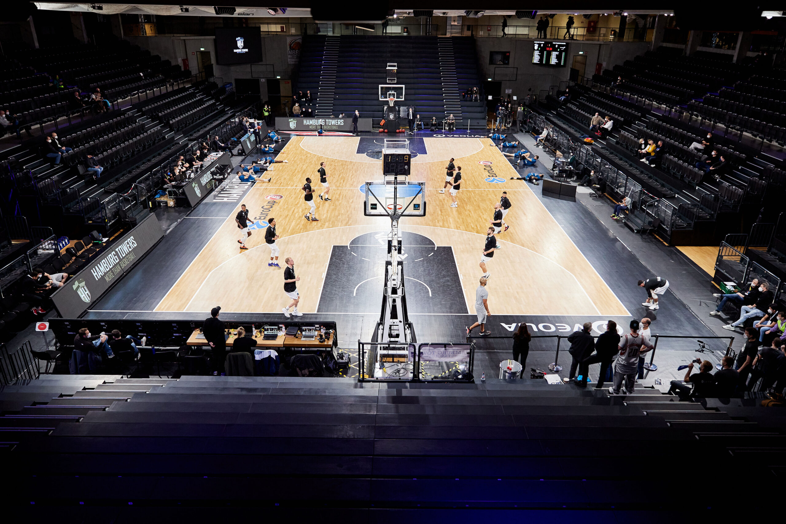 Leeres Basketballfeld in einer Halle mit wenigen Spielern, die sich aufwärmen, und leeren Tribünen