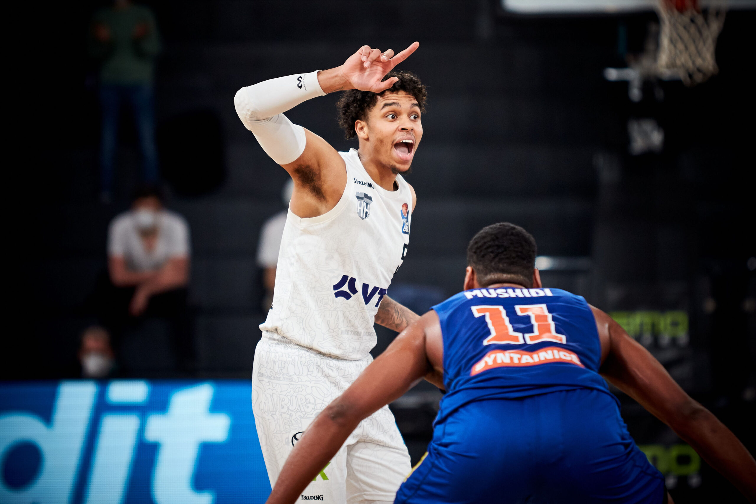 Basketballspieler in weißer Uniform gestikuliert und spricht energisch, verteidigt gegen Spieler in blauer Nummer 11