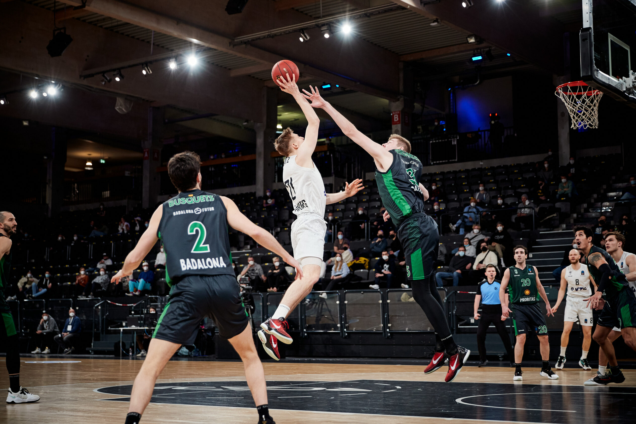 Basketballspieler in weiß springen zum Korb, während zwei Spieler in schwarzen Trikots mit grünen Akzenten verteidigen, Zuschauer im Hintergrund