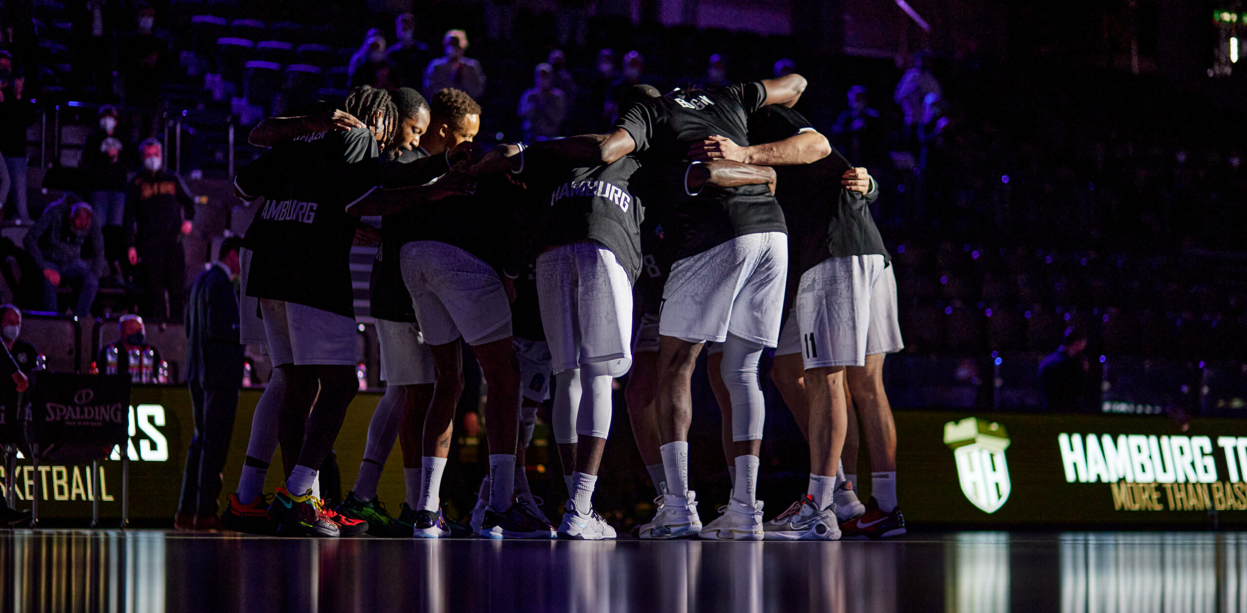 Basketballteam in schwarzer Trainingskleidung steht im Kreis und umarmt sich vor dem Spiel in einer dunklen Halle