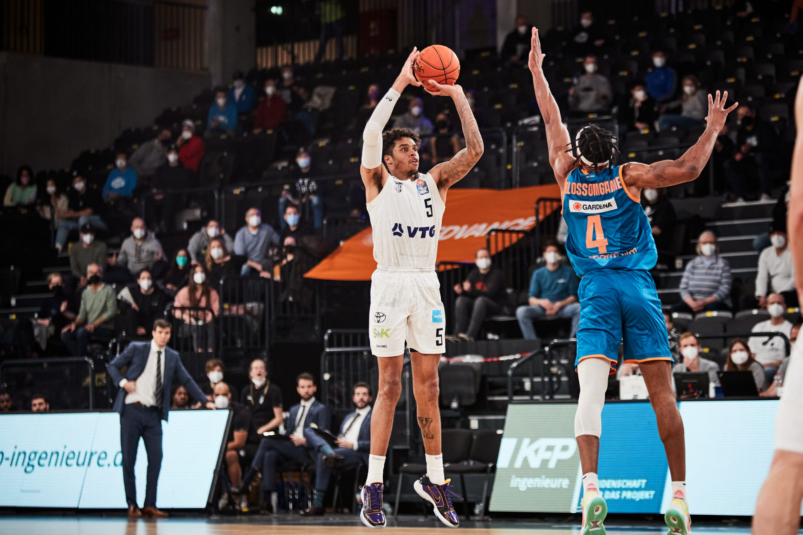 Basketballspieler in weißer Uniform wirft den Ball, während ein Spieler in blauer Uniform mit erhobenen Armen verteidigt, Zuschauer im Hintergrund
