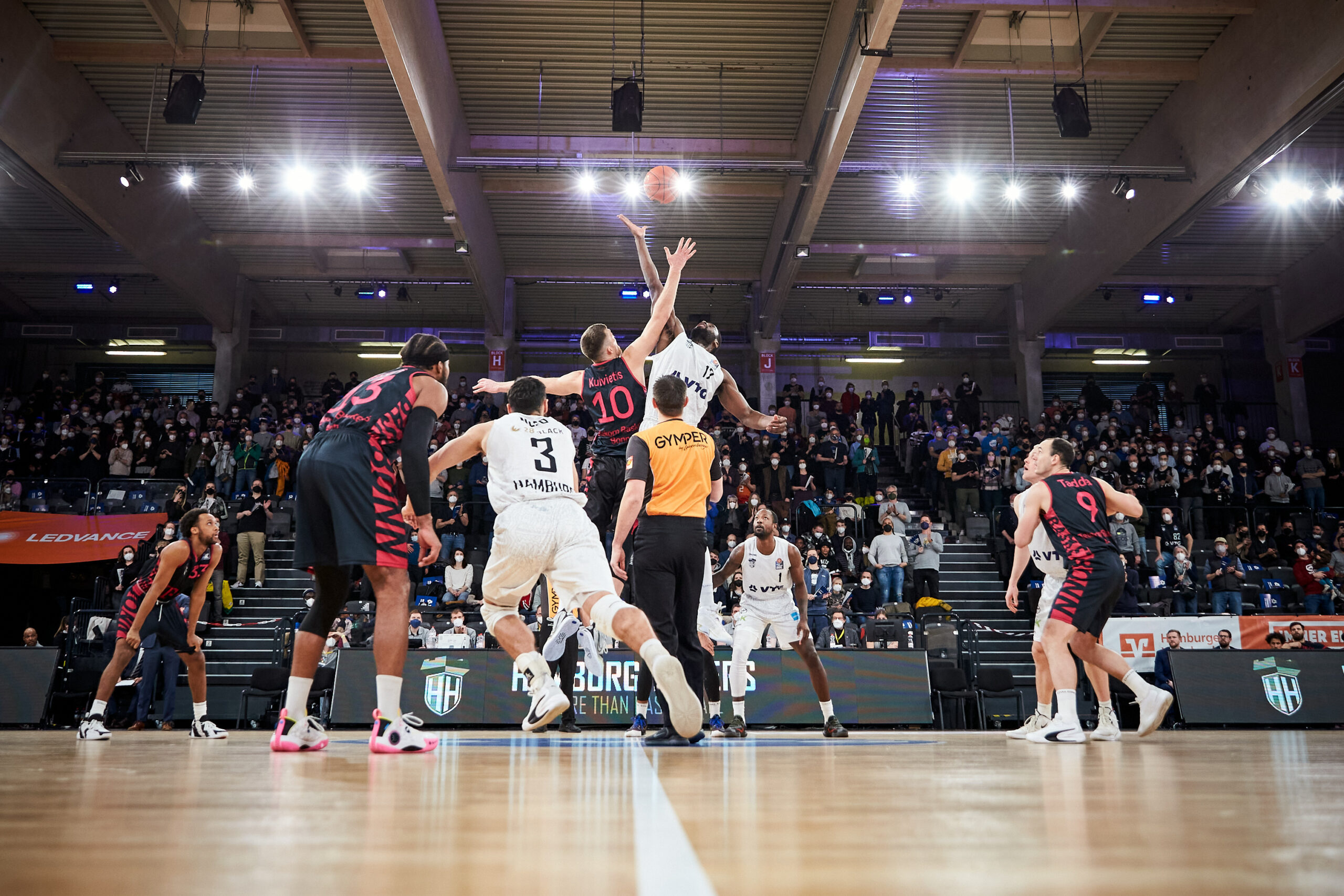 Basketballspiel beim Sprungball zwischen zwei Spielern in einer Halle mit Zuschauern auf den Tribünen