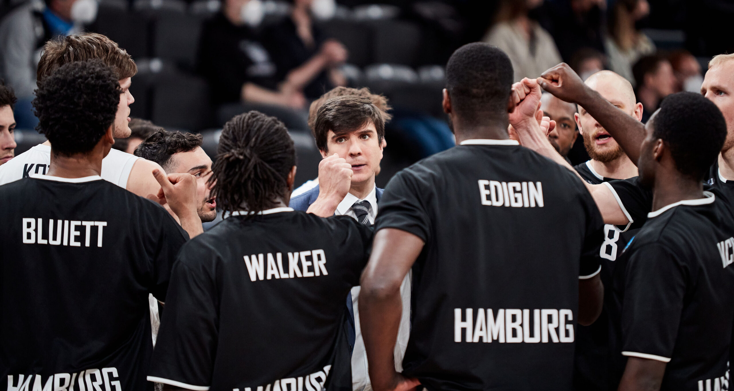 Basketballteam Hamburg in schwarzen Trikots versammelt sich um den Trainer, der Anweisungen gibt.