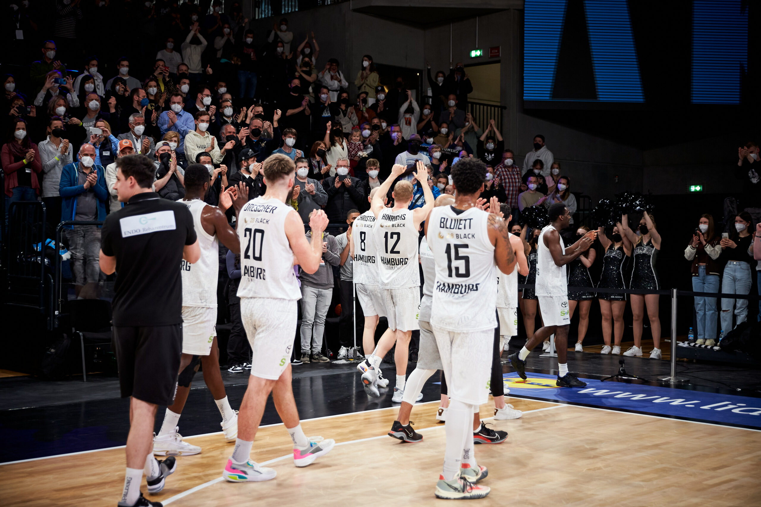 Basketballspieler in weißen Trikots applaudieren vor jubelndem Publikum in einer Halle