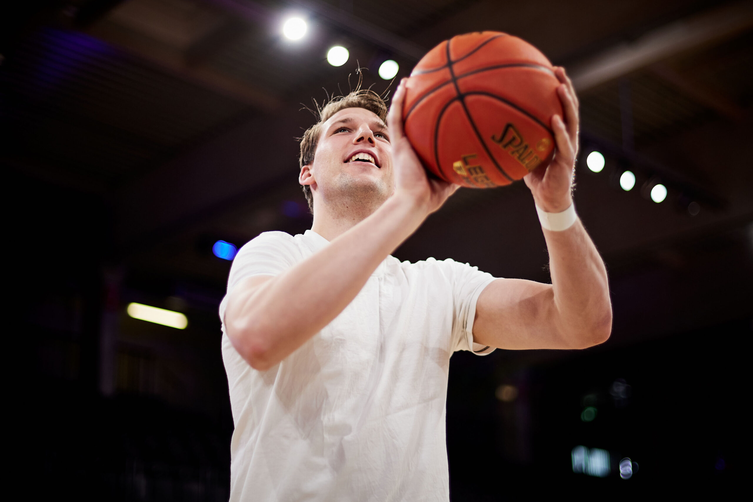Mann in weißem T-Shirt hält Basketball zum Wurf in einer Halle mit Deckenbeleuchtung