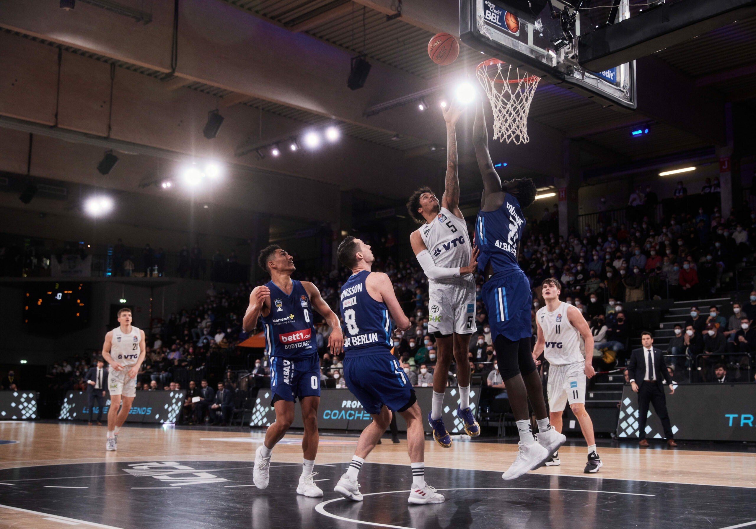 Basketballspieler in weißem Trikot versucht Korb zu erzielen, während Spieler in blauen Trikots verteidigen, Zuschauer im Hintergrund