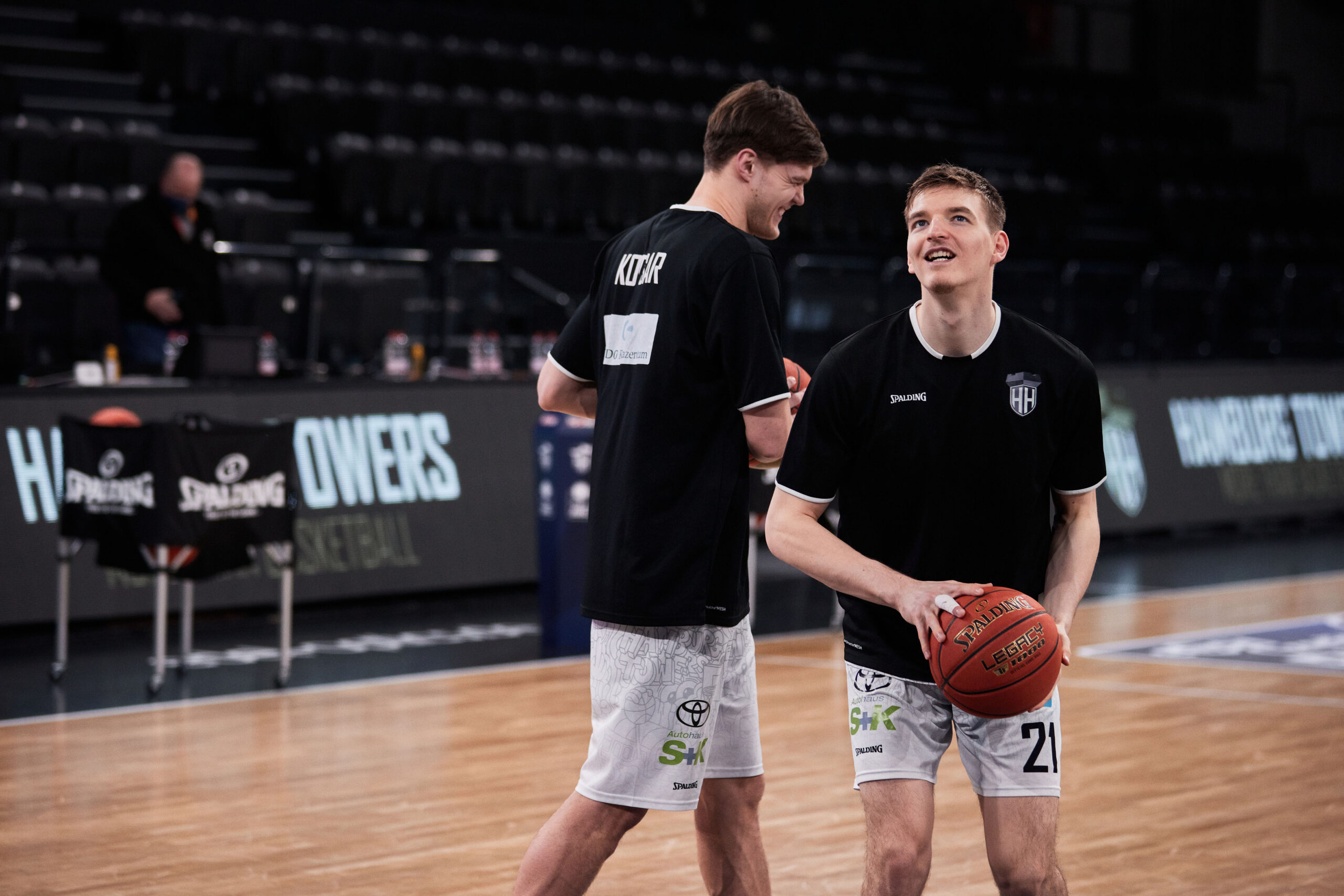 Zwei Basketballspieler in schwarzen Trikots und weißen Shorts auf dem Spielfeld, einer hält einen Basketball und lächelt.
