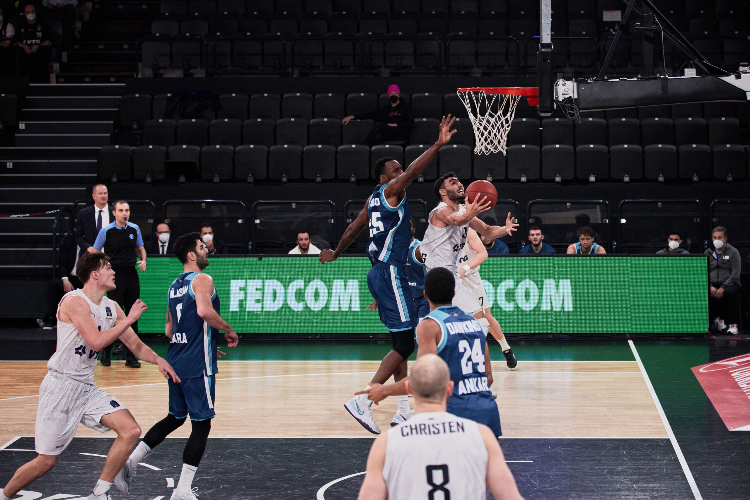 Basketballspieler in weißer Trikotfarbe versucht Korb zu erzielen, während Spieler in blauen Trikots verteidigen, Spiel findet in Halle mit leeren Rängen statt