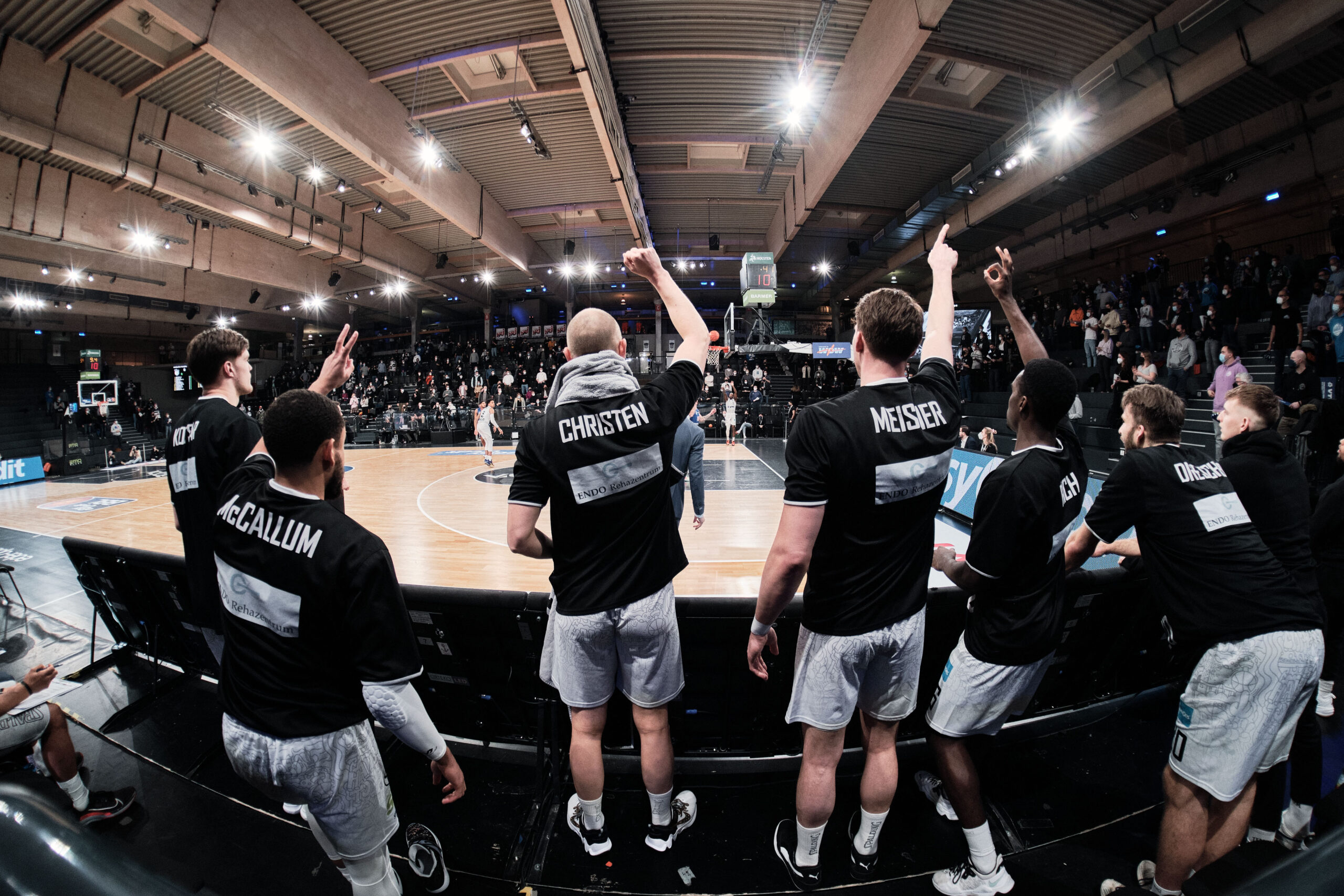 Basketballspieler auf der Bank heben jubelnd die Arme während eines Spiels in einer Halle mit Zuschauern