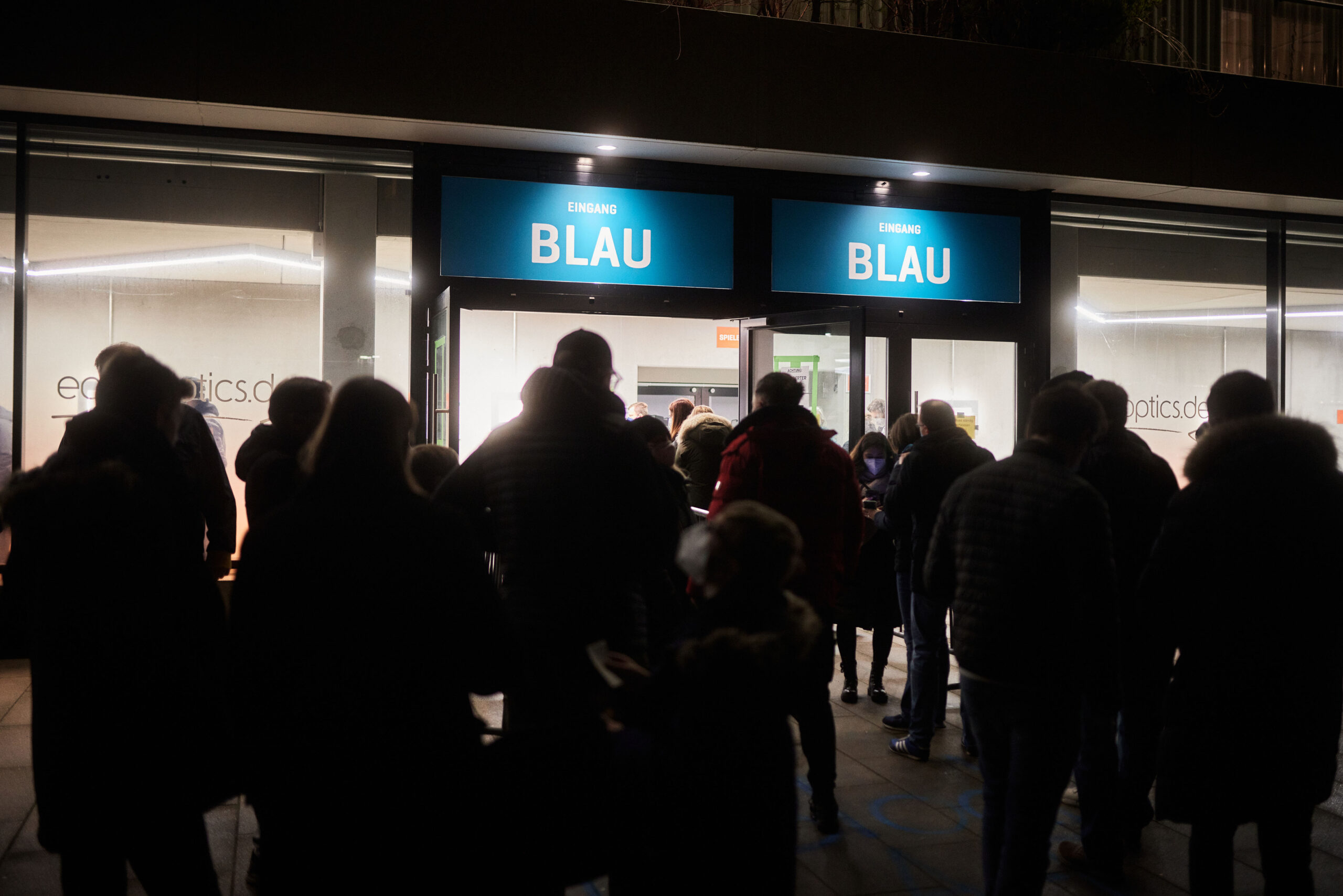 Menschenmenge betritt abends durch zwei Eingänge mit blauen Schildern und der Aufschrift 'BLAU' ein Gebäude
