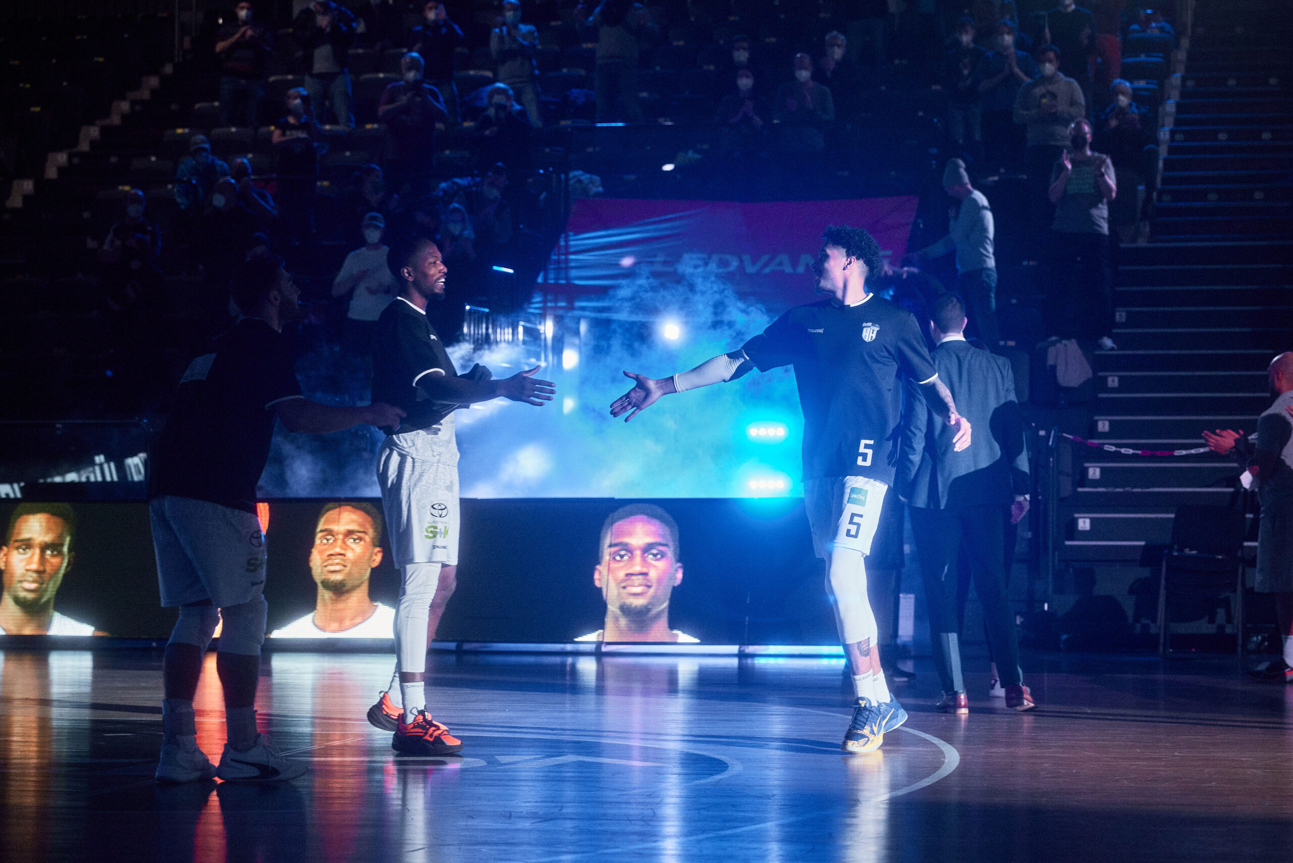 Zwei Basketballspieler in dunklen Trikots geben sich vor einem Publikum in einer Halle die Hand, beleuchtet von Bühnenlicht und mit großen Spielerporträts im Hintergrund