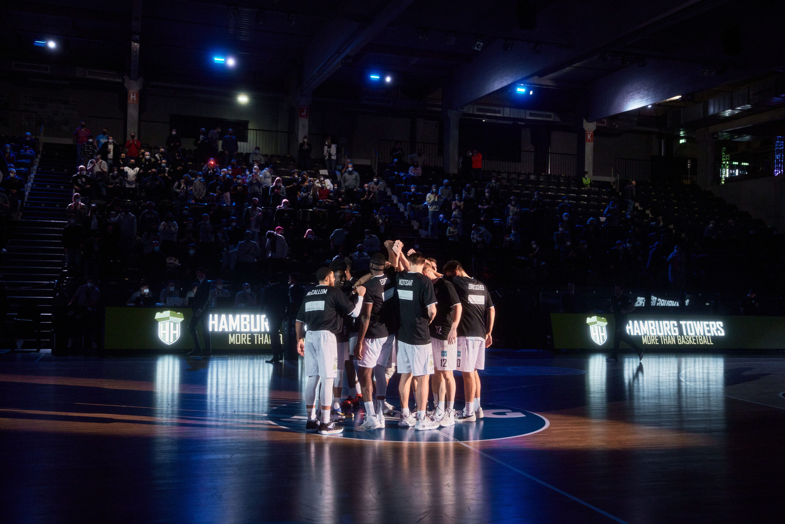 Basketballteam in weißen Shorts und schwarzen Trikots steht im Kreis auf dem Spielfeld, umgeben von dunkler Halle und Zuschauern im Hintergrund