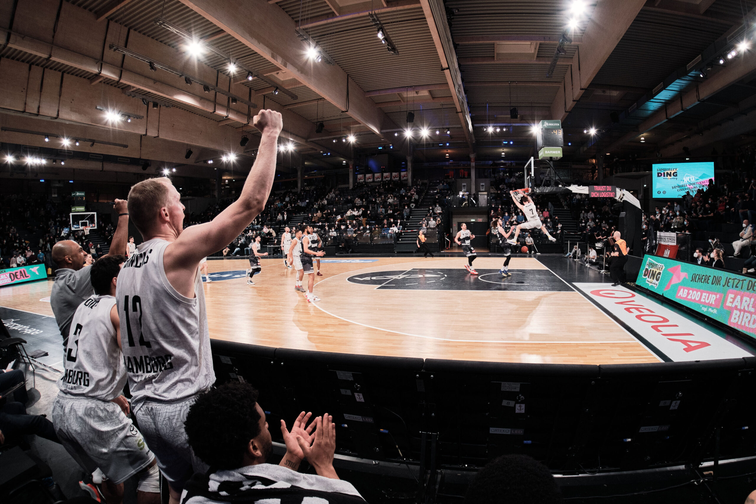 Basketballspieler in weißen Trikots jubeln auf der Bank, während ein Spieler einen Dunk am Korb ausführt in einer Halle mit Zuschauern