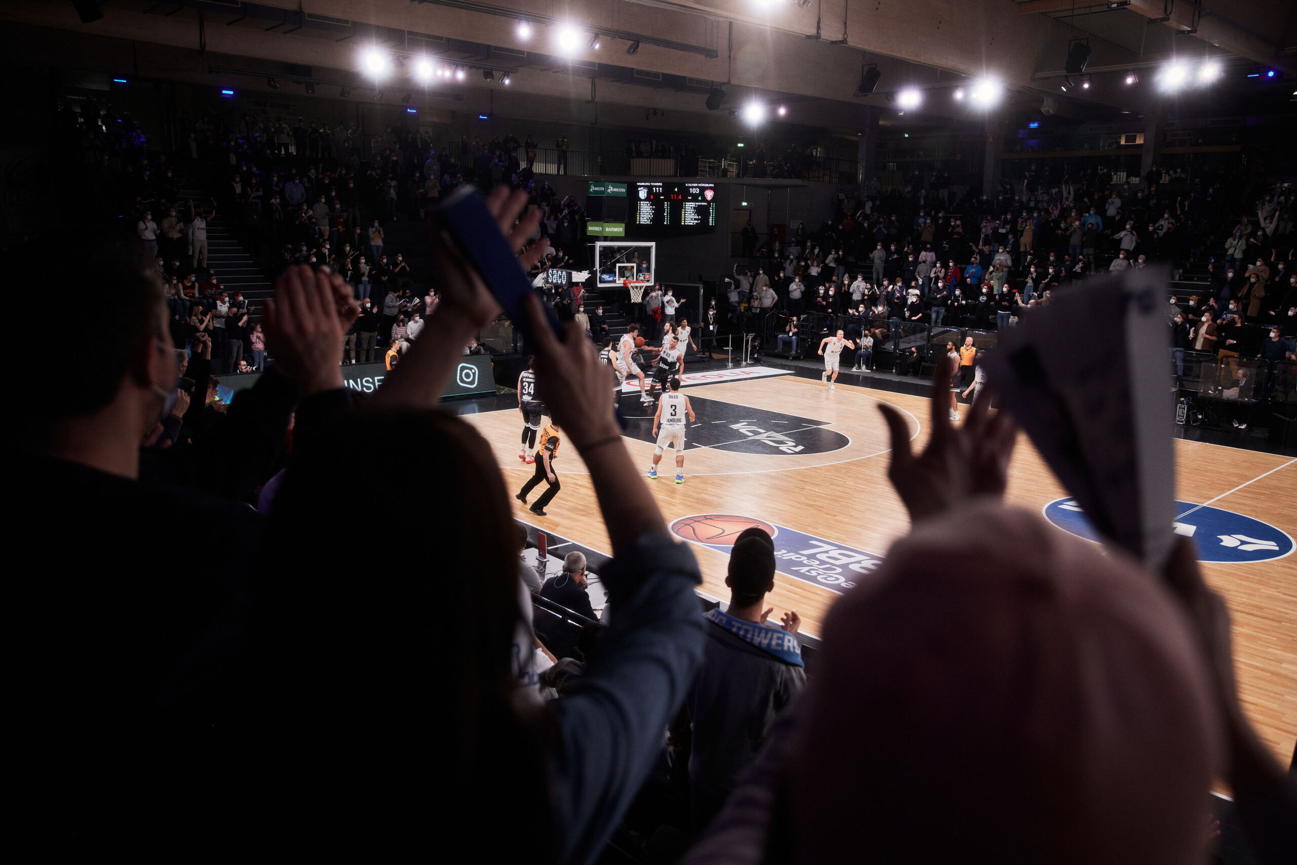 Zuschauer jubeln in einer Halle während eines Basketballspiels mit Spielern auf dem Spielfeld und beleuchtetem Spielfeld