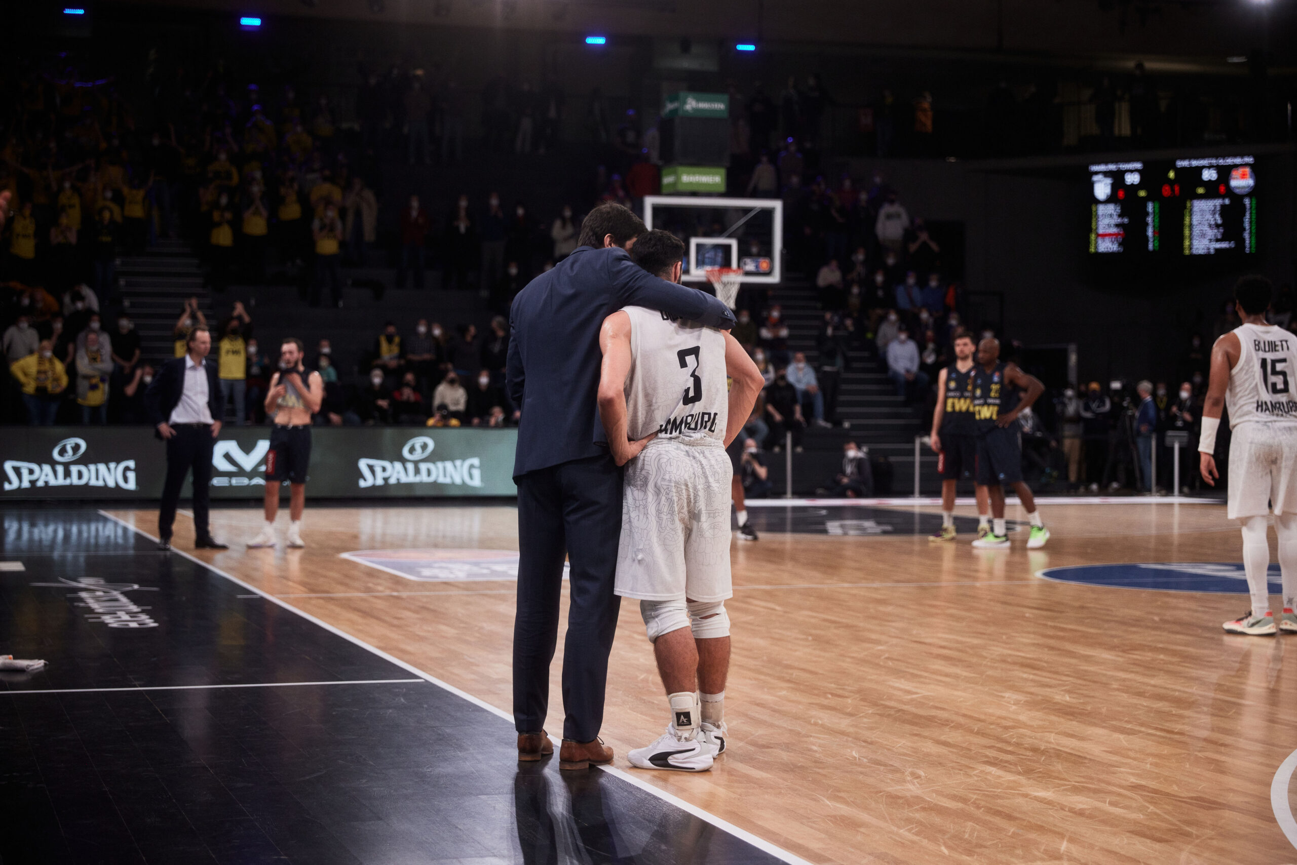 Basketballspieler in weißem Trikot mit Nummer 3 wird von einem Mann im Anzug auf dem Spielfeld getröstet, Zuschauer und andere Spieler im Hintergrund