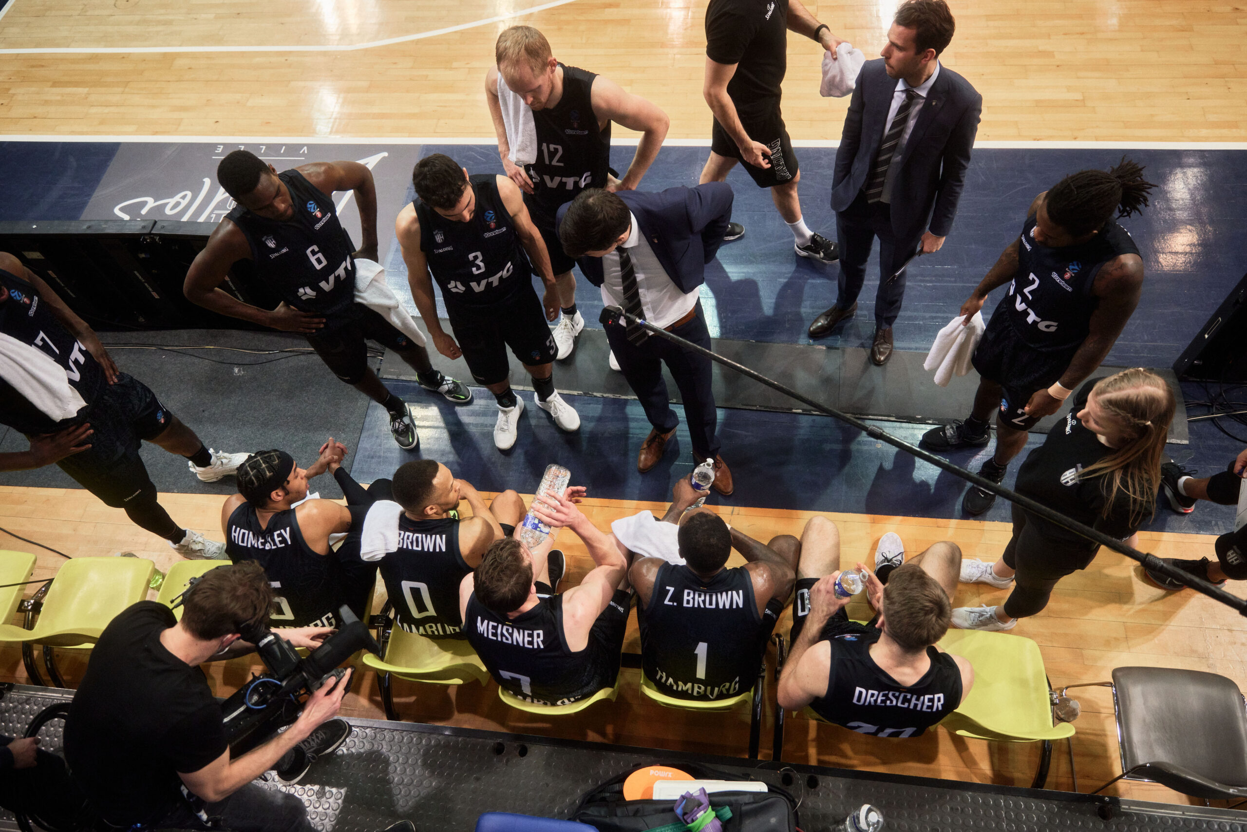 Basketballteam in schwarzen Trikots während einer Auszeit, Trainer gibt Anweisungen am Spielfeldrand