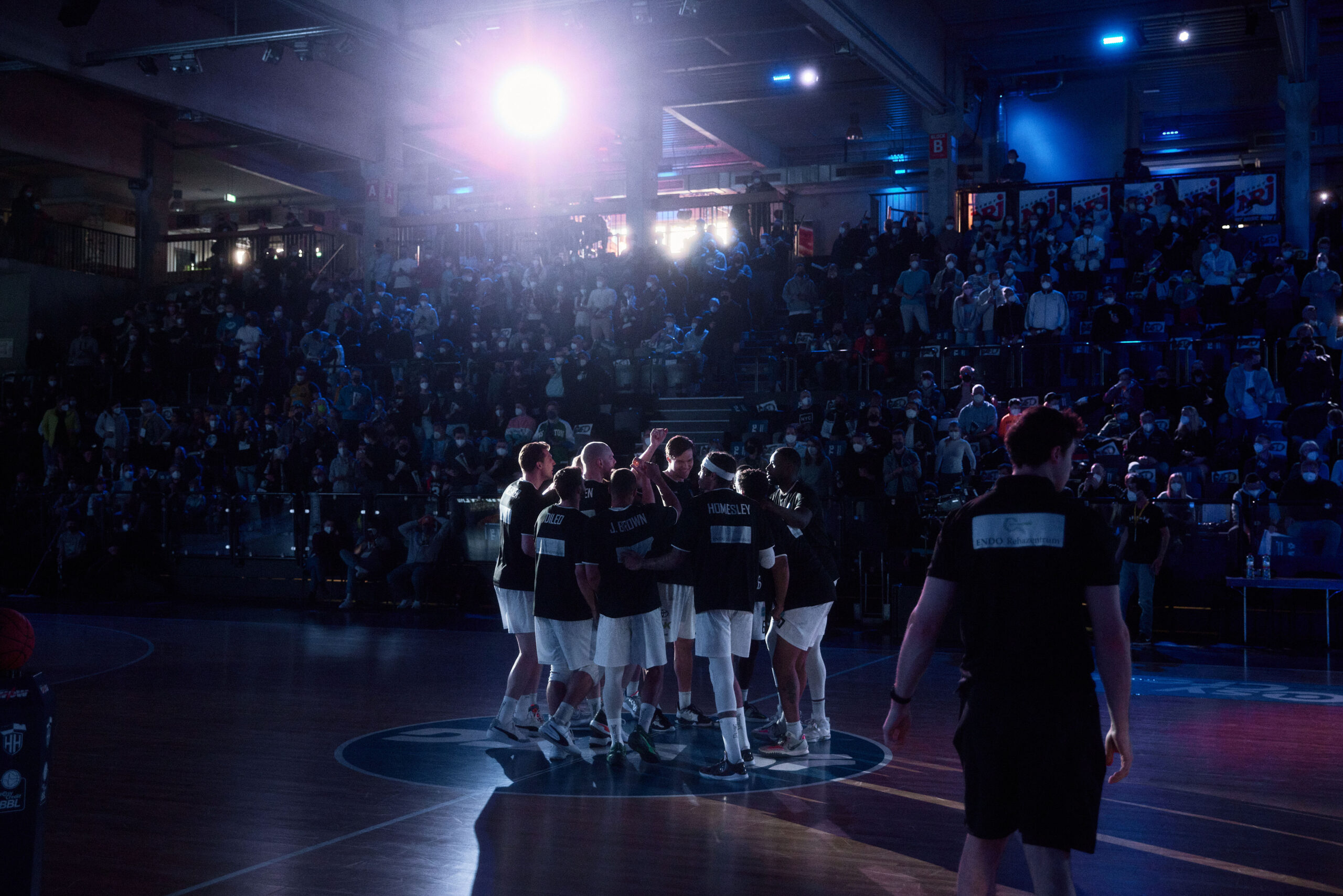 Basketballspieler in einem Kreis auf dem Spielfeld, umgeben von Zuschauern in einer dunklen Halle mit Bühnenlicht