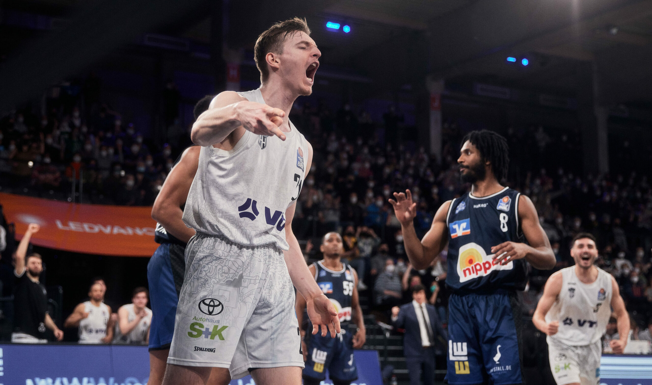 Basketballspieler in weißem Trikot schreit jubelnd, während Spieler in dunklen Trikots zuschauen, Spiel in Halle mit Zuschauern